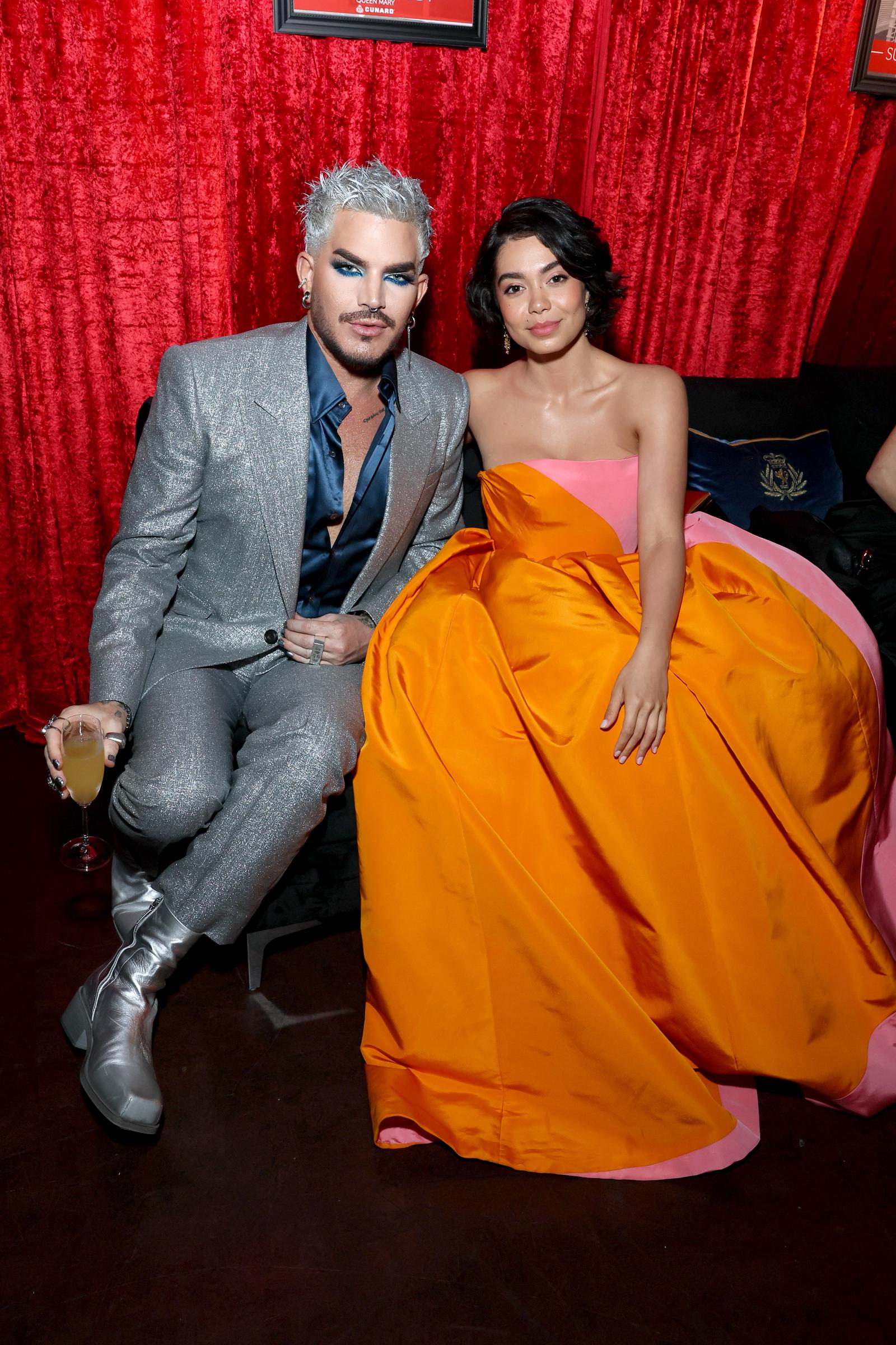Seated beside Auli'i Cravalho on a velvet sofa, Adam Lambert relaxes with a drink in hand. His shimmering silver look contrasts with her bold orange-pink gown, creating a luxe, intimate image that reflects the night's fashionable highs.