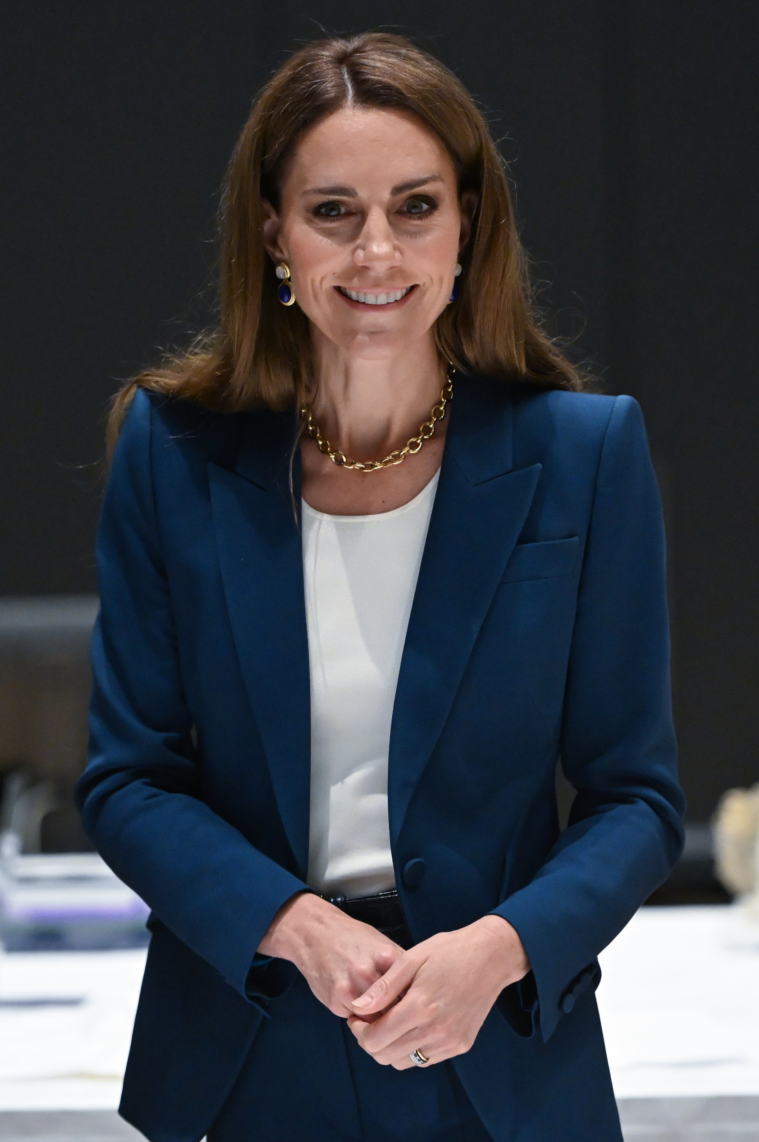 Princess Catherine smiles softly in a tailored blue suit and white top. Her posture is upright, hands gently clasped, radiating calm confidence as she stands inside the V&A East Storehouse.