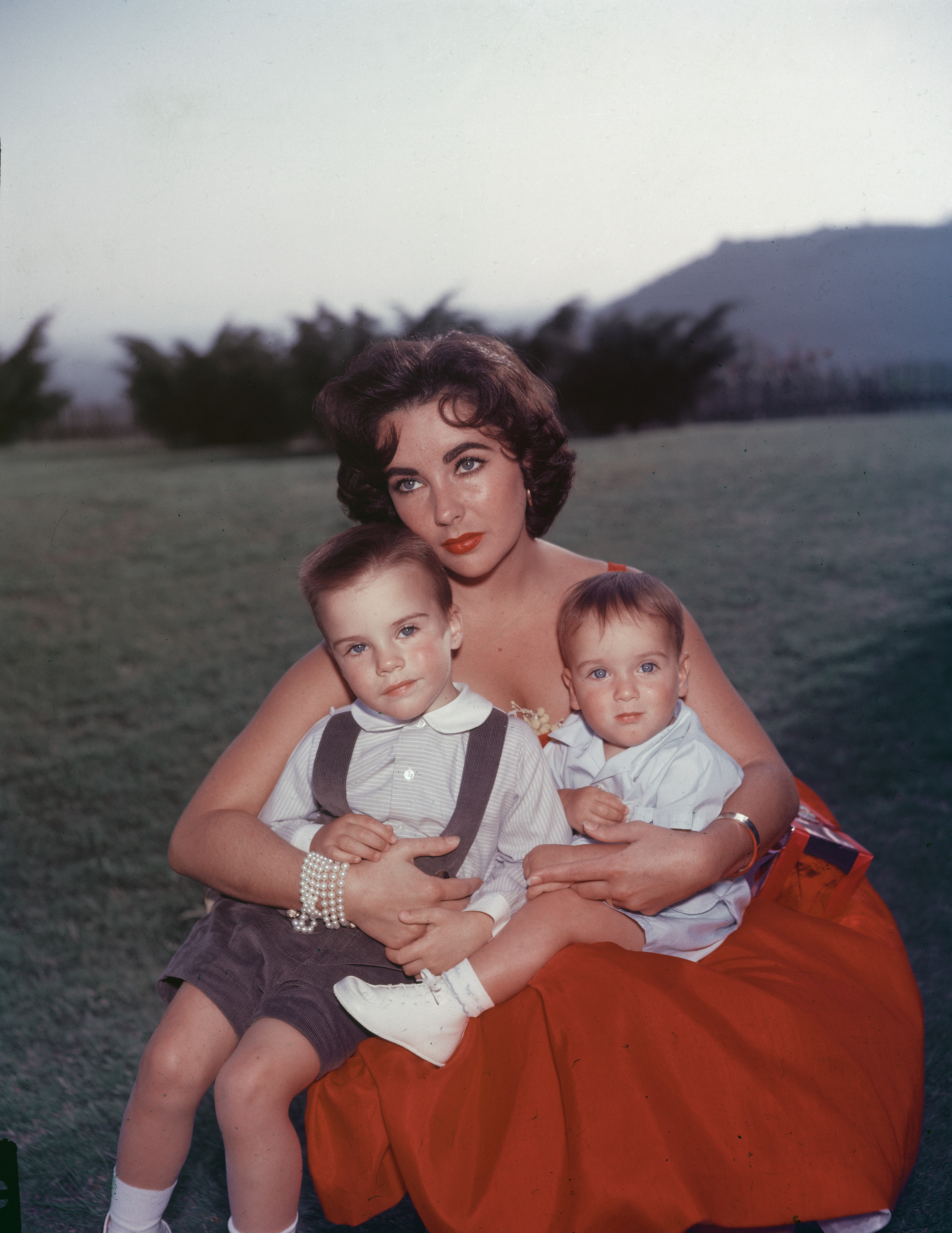 Elizabeth Taylor sentada en un vestido de noche rojo con sus hijos Michael y Christopher Wilding en su regazo | Foto: Getty Images