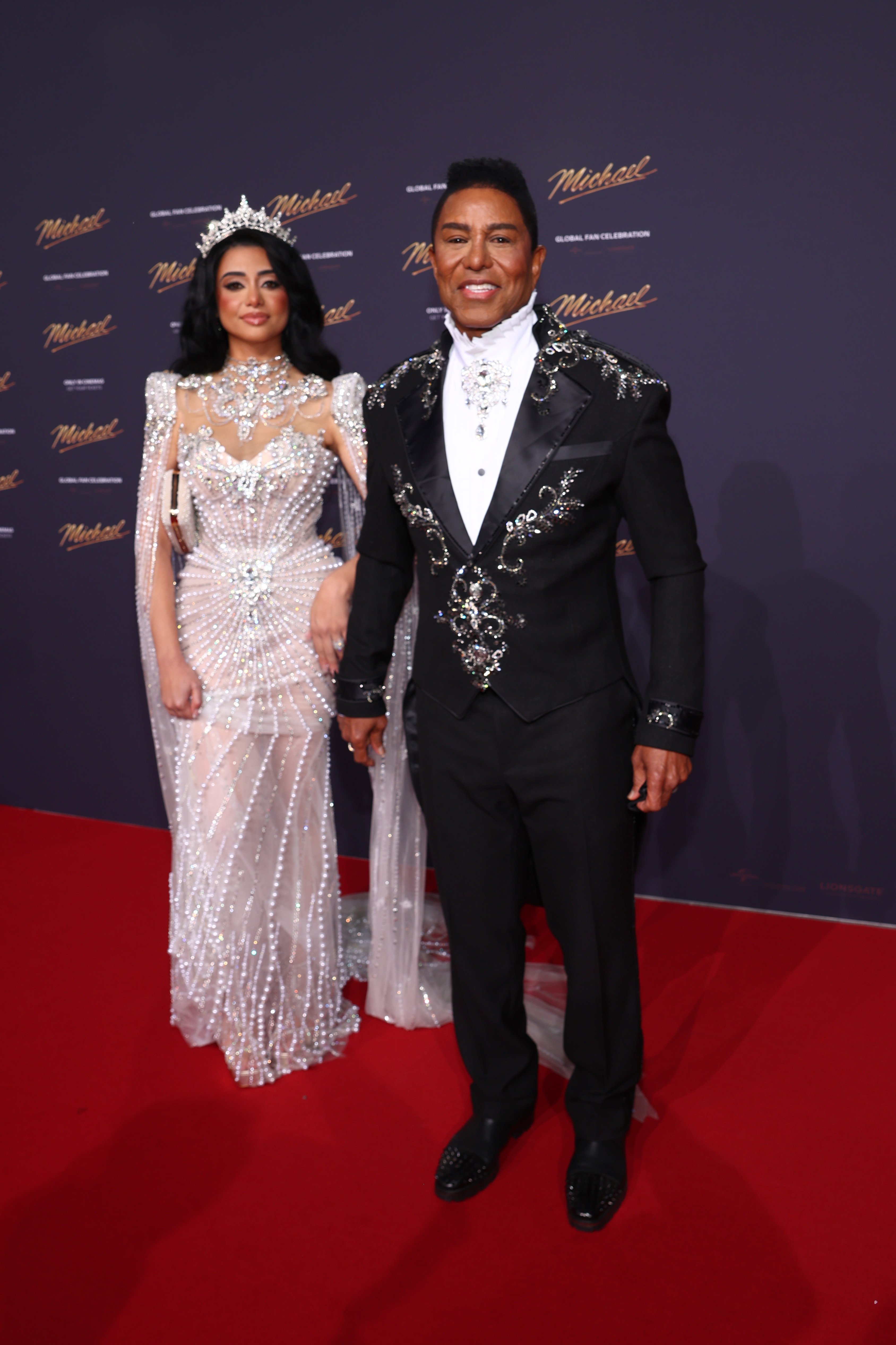 Jermaine Jackson poses alongside his wife, Maday, on the red carpet, maintaining his signature polished style in the embellished black ensemble. With the event backdrop glowing behind them, his poised stance and subtle smile reinforce a composed, red carpet-ready presence.