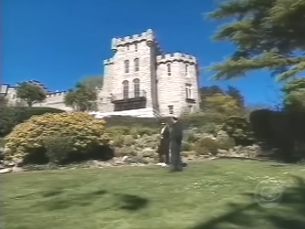 Set against a cloudless blue sky, the castle rises above manicured lawns and flowering shrubs, its turrets and arched windows evoking a romantic, almost storybook quality. The carefully tended grounds soften the fortress-like façade, revealing the balance between grandeur and domestic calm. | Source: YouTube/AndreW