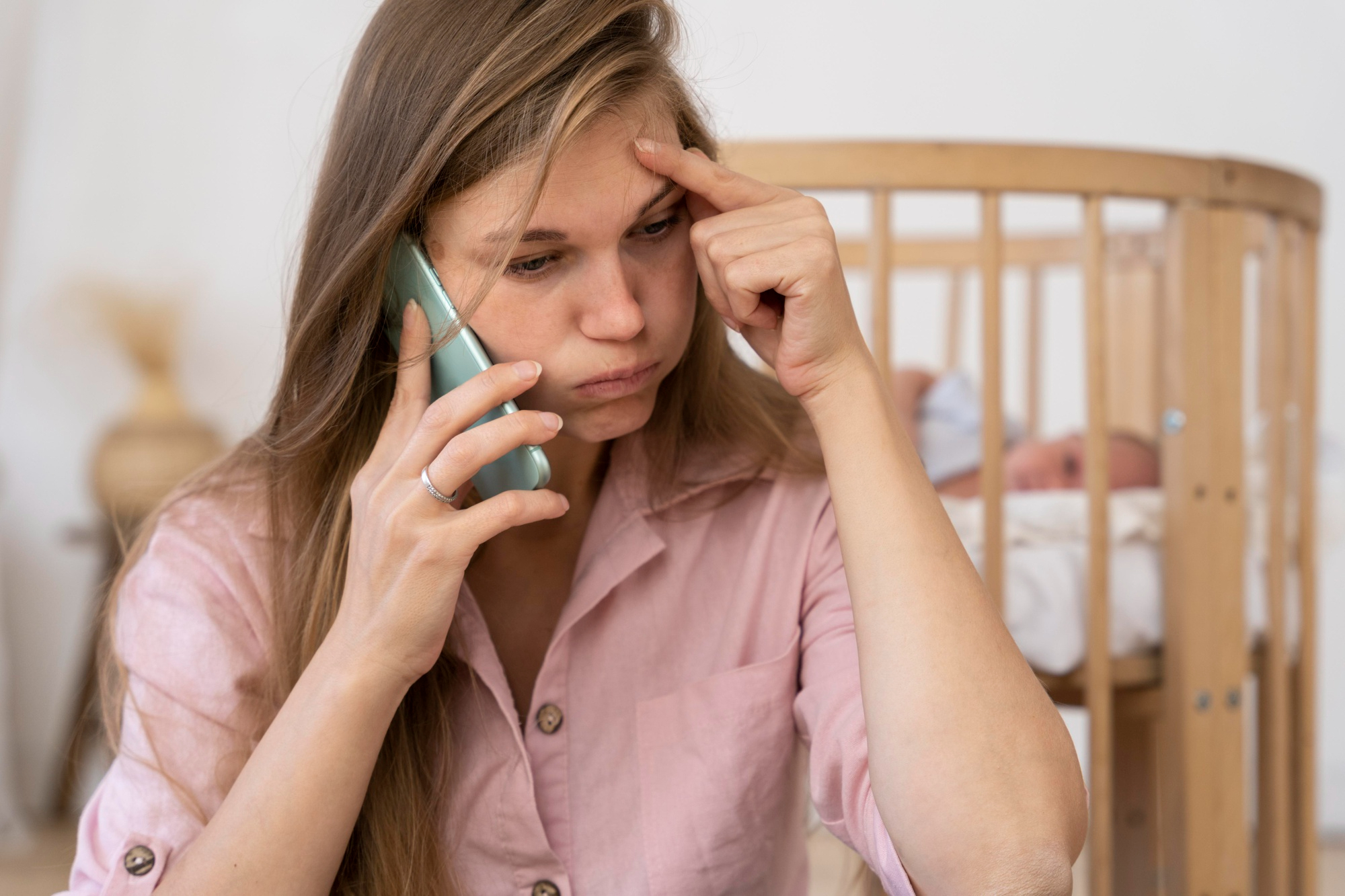 A frustrated woman talking on the phone with her baby lying in a cot behind her | Source: Freepik