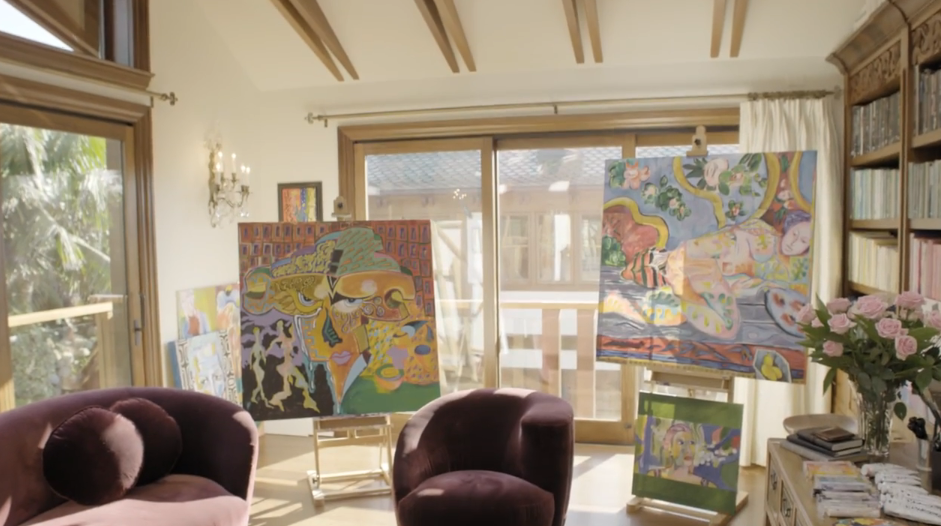 Vibrant abstract paintings rest on easels throughout Pierce Brosnan's art studio, illuminated by bright sunlight streaming in through vaulted ceilings and sliding glass doors. Plush plum velvet armchairs offer a comfortable spot for inspiration, while built-in wooden bookshelves line the adjacent wall beside a vase of pink roses. | Source: YouTube/Architectural Digest