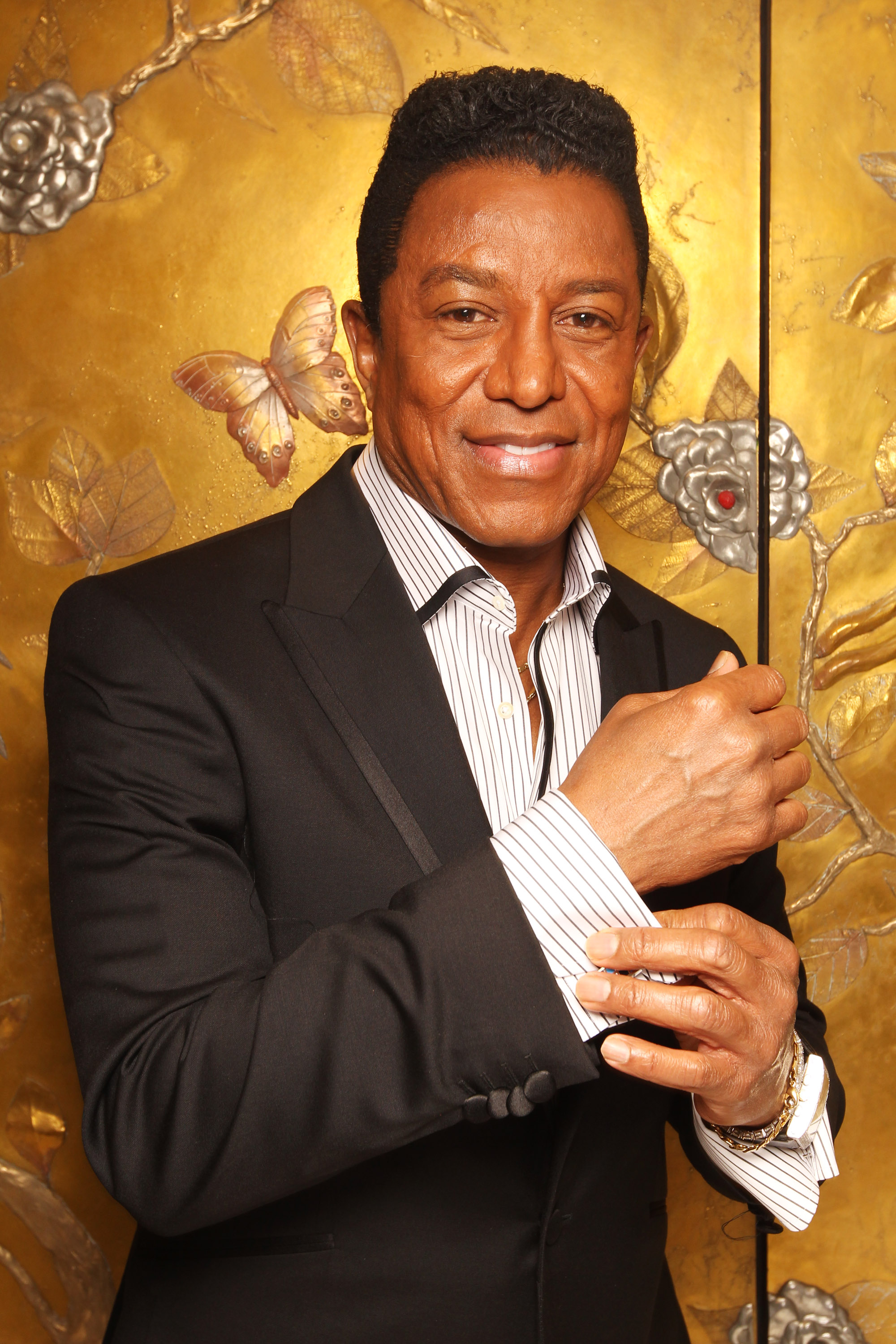Jermaine Jackson poses for a portrait at The Dorchester in London, adjusting his cufflinks in a sharply tailored black suit. Set against a gold-embossed backdrop, the refined styling and poised gesture emphasize his polished, timeless elegance.