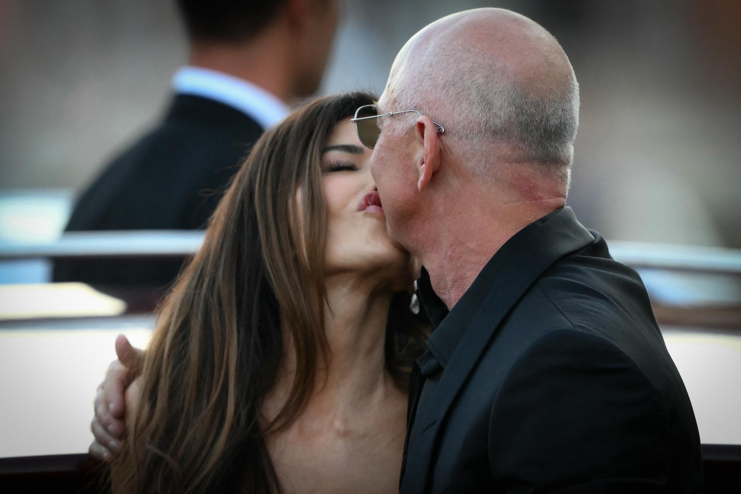 Lauren Sánchez and Jeff Bezos share a kiss as a suited guard looks on, the candid moment set against blurred buildings and the evening light reflecting off the water.