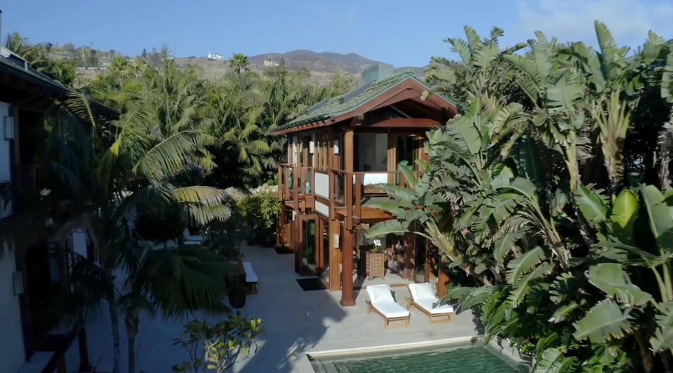 Nestled amidst dense tropical foliage and towering banana palms, a two-story wooden pool house features an open-air upper balcony and ground-level glass doors opening onto the poolside patio. White lounge chairs line the edge of the sparkling pool, while the surrounding greenery frames a peaceful view toward the Malibu hills behind the estate. | Source: YouTube/Architectural Digest