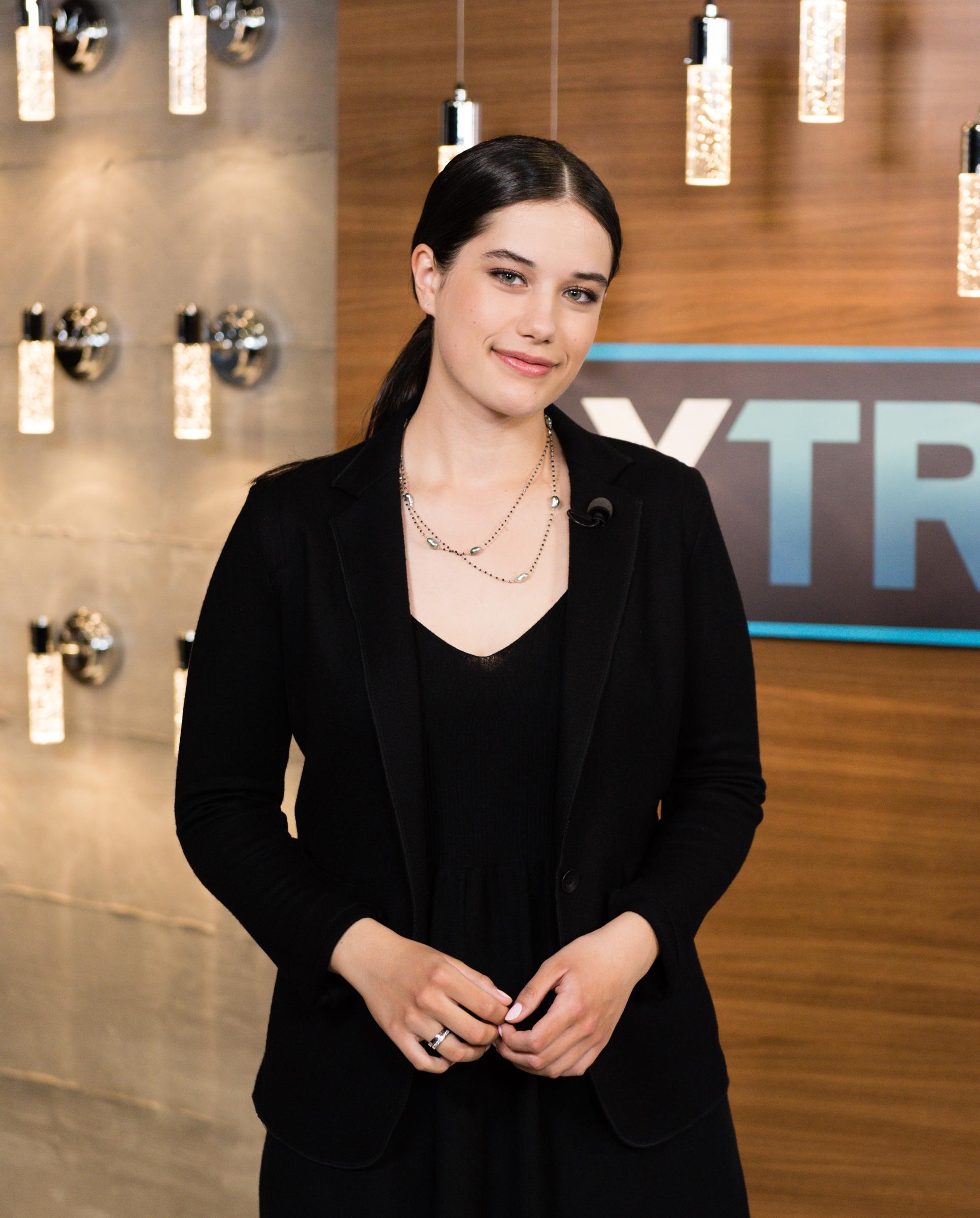Ella Bleu Travolta smiles softly in a black blazer and layered necklace during a 2019 visit to "Extra" at Universal Studios Hollywood. The studio appearance captured her in a sleek, understated look.