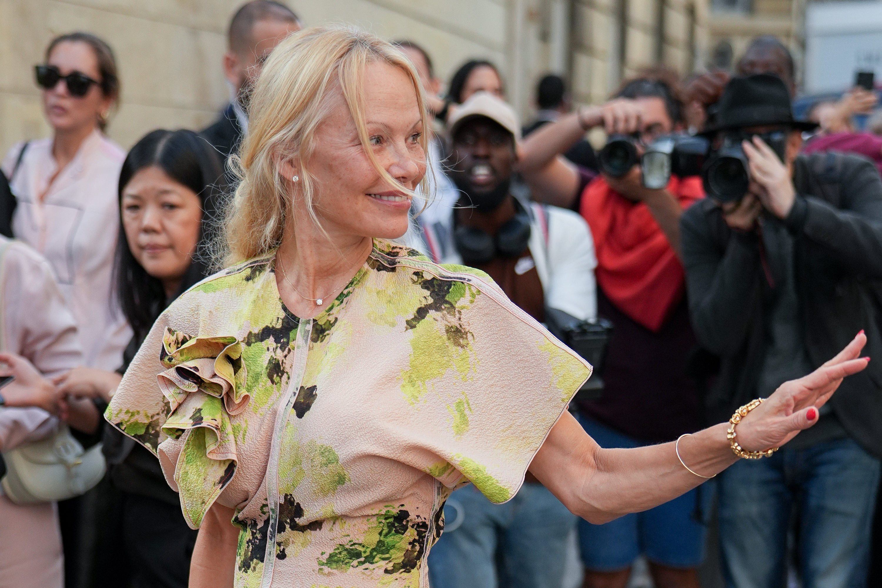 Pamela Anderson, with her blonde hair pulled back loosely and a few strands escaping to frame her face, wears a pale pink and green floral-patterned dress with ruffled details on the sleeve and a gold bracelet on her wrist. She appears to be without makeup as she attends the Victoria Beckham Womenswear Spring/Summer show as part of Paris Fashion Week on September 29, 2023, in Paris, France.