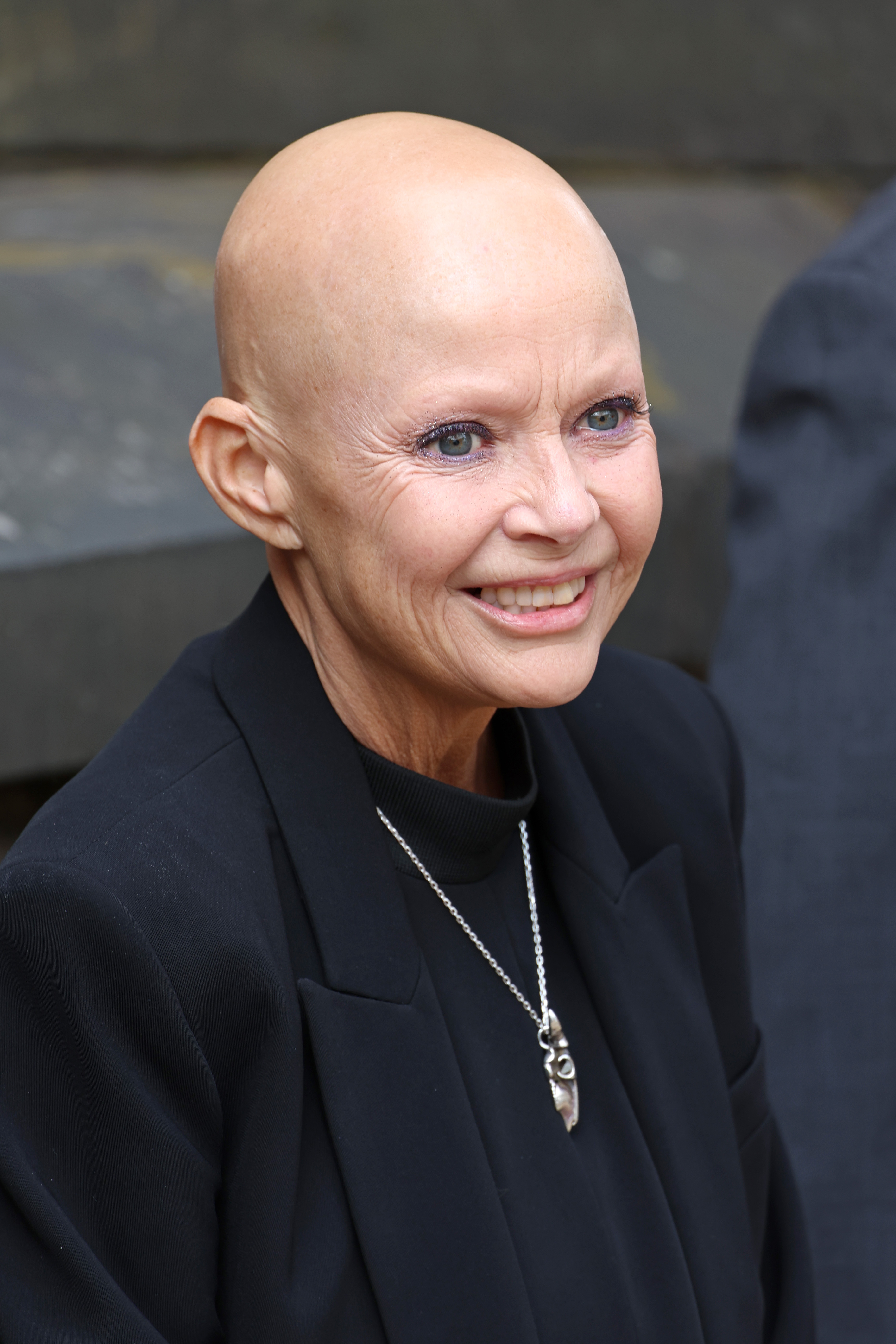 A close outdoor portrait of Porter, her brow-less face framed by delicate lilac eyeshadow and a fine silver pendant necklace, deep laughter lines crinkling around her eyes as she smiles widely. Pictured at a celebration marking the 900th anniversary of the City of Edinburgh at Edinburgh Castle, July 2024.