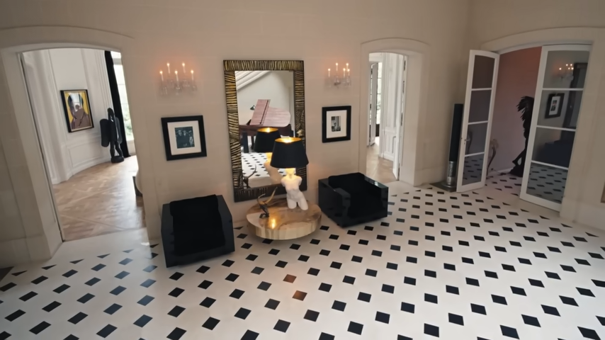 The elegant second-floor landing features a textured gold-framed mirror, a sculptural torso lamp on a polished stone table, and low-slung black armchairs flanked by arched doorways. | Source: YouTube/@Archdigest