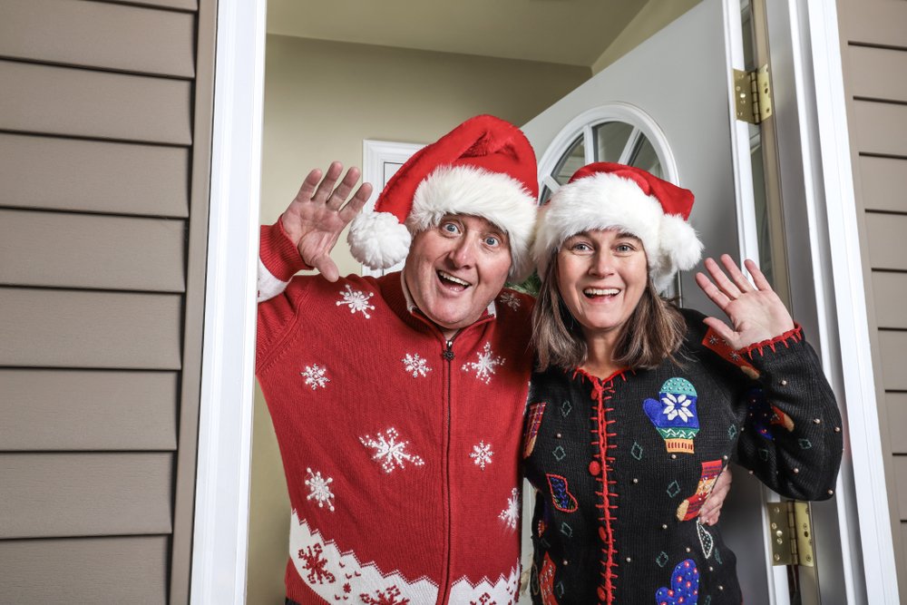 Pareja saludando desde la puerta de su hogar vistiendo suéteres navideños. | Foto: Shutterstock