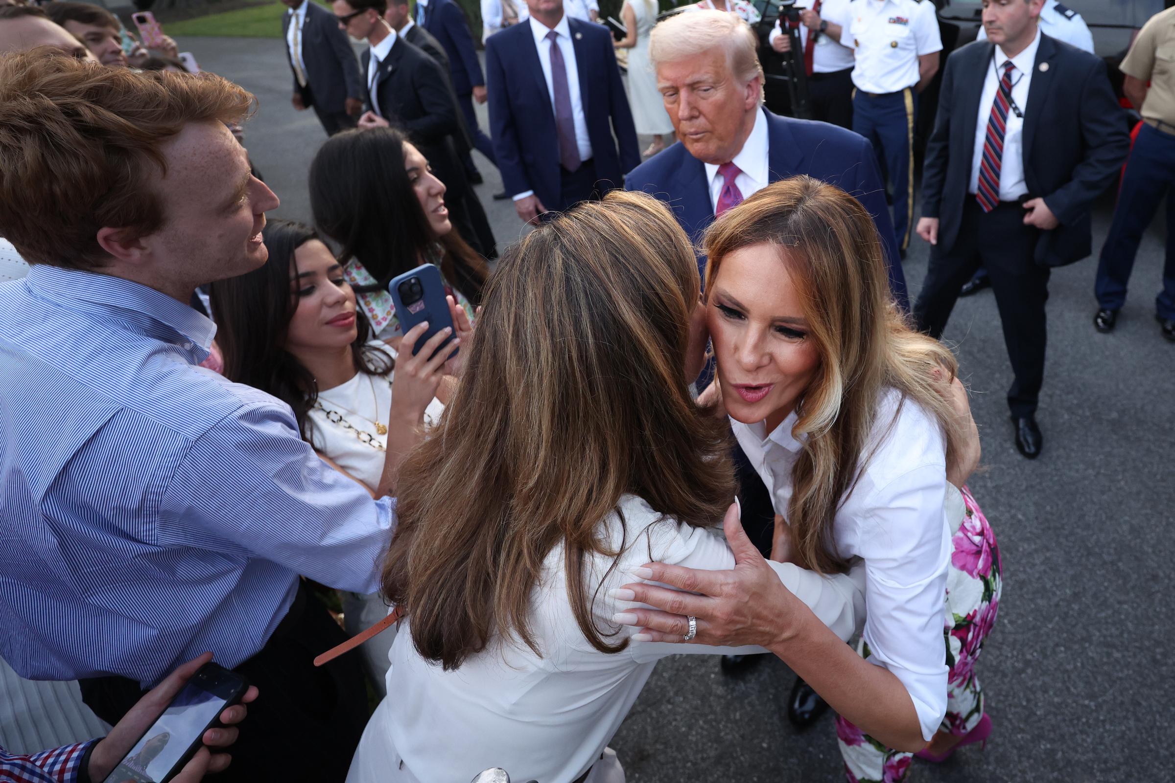 Melania Trump embraces Rep. Maria Elvira Salazar during the 2025 Congressional Picnic at the White House. As attendees look on and take photos, Donald Trump stands nearby, observing the warm exchange amid a lively crowd and security presence.