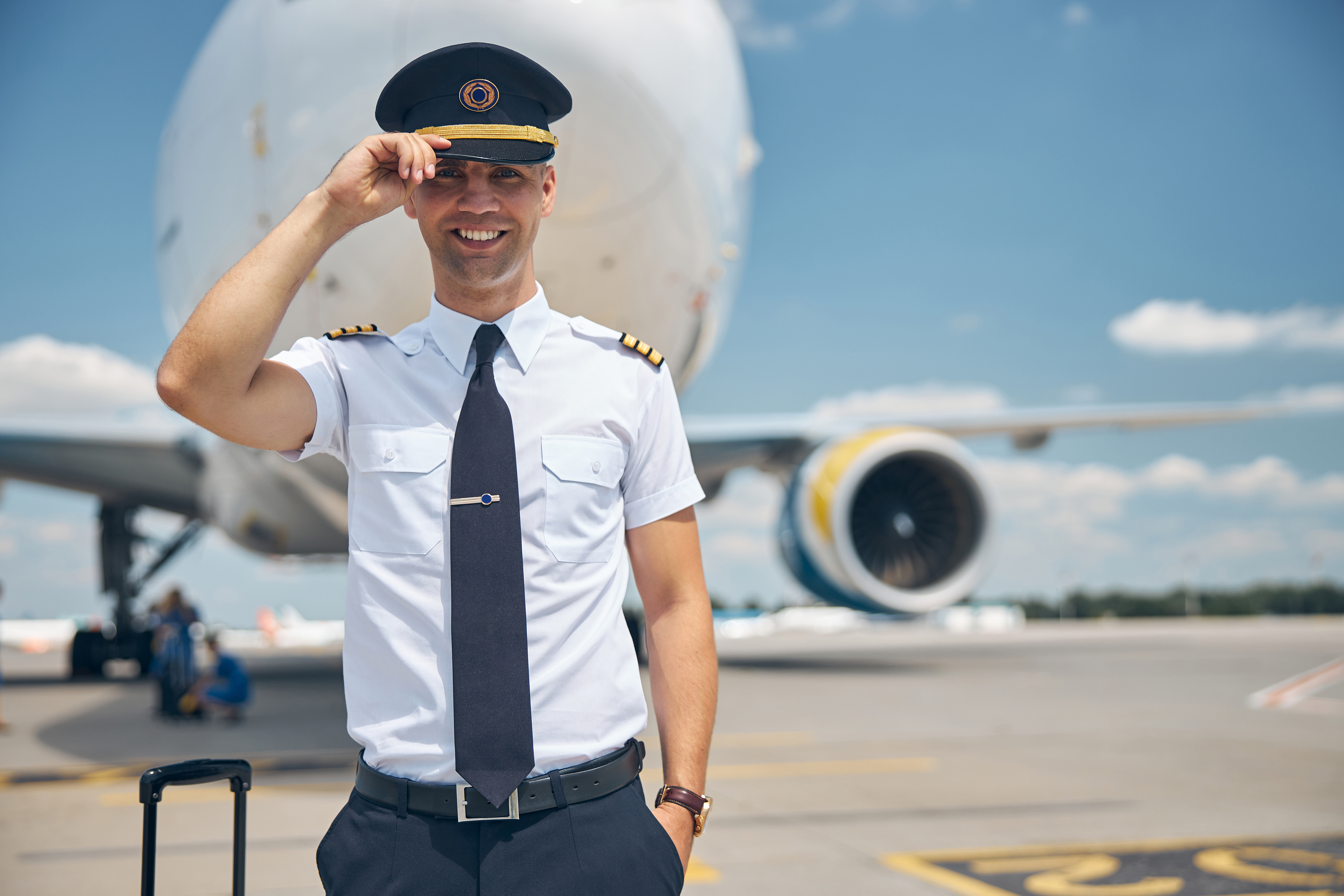 Cheerful young man airline worker touching captain hat and smiling while standing in airfield with airplane on background. | Source: Shutterstock