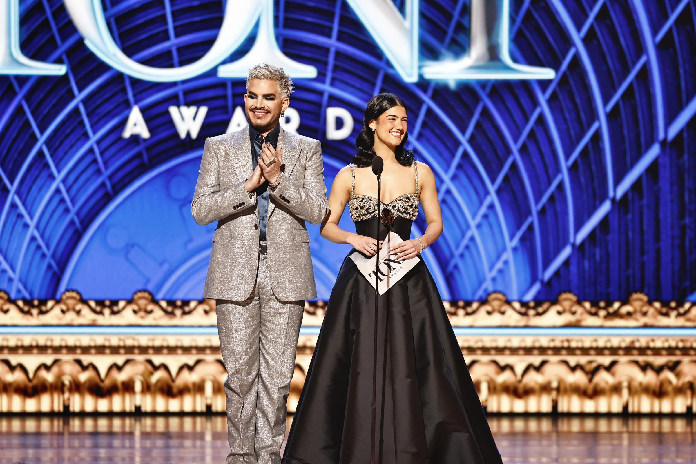 Adam Lambert and Charli D'Amelio stand side-by-side on stage, both beaming as they prepare to present. His metallic suit glistens against the deep blue backdrop, while her strapless gown adds contrast to the vibrant stage palette.