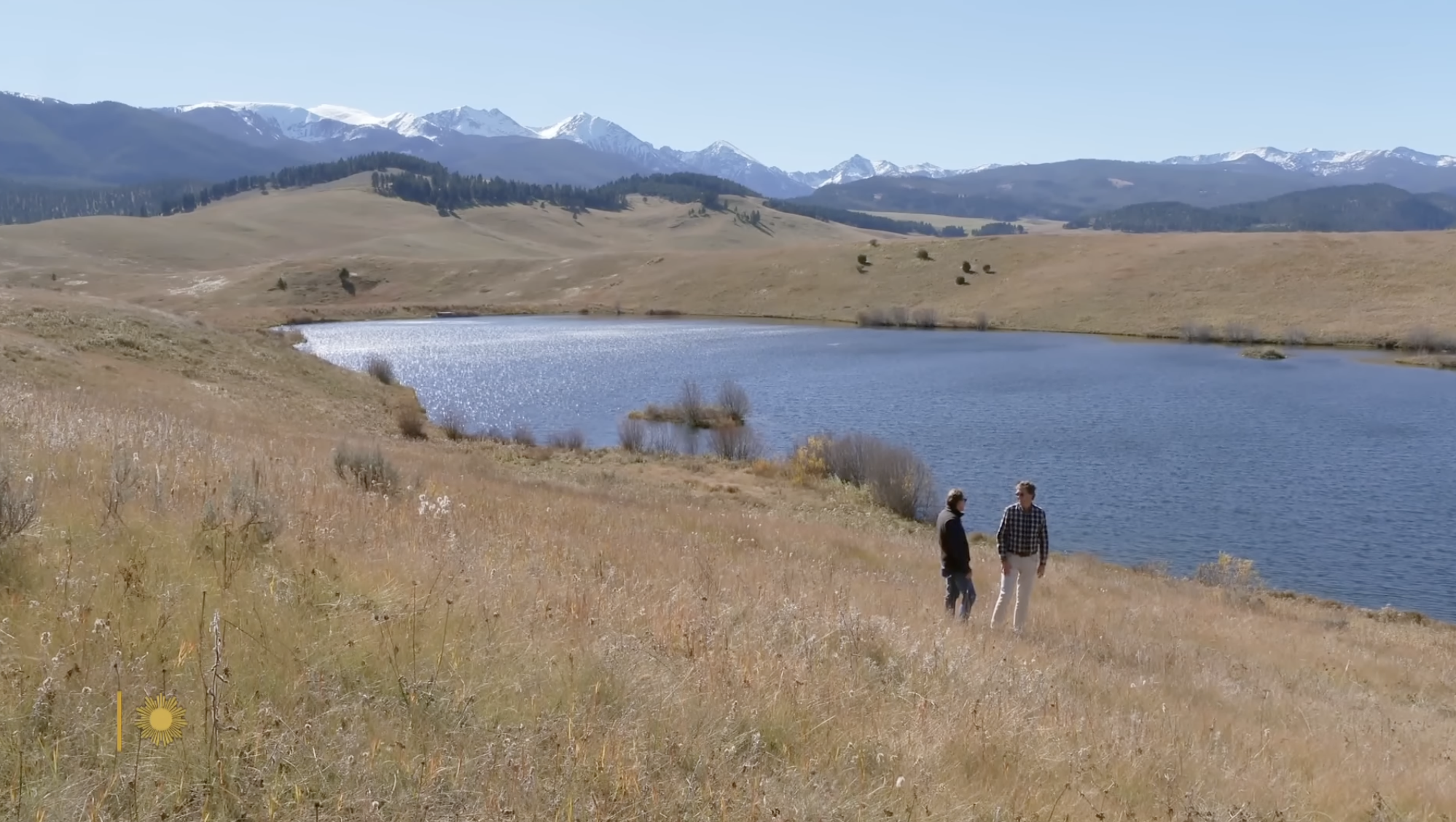 Two people stand beside a tranquil lake surrounded by rolling plains and mountain scenery on one of Ted Turner’s expansive wilderness properties. | Source: YouTube/CBS Sunday Morning