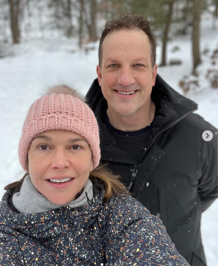 Sutton Foster and Ted Griffin pose for a winter selfie on a snowy trail surrounded by trees. Sutton stays cozy in a pink knit hat and patterned jacket, while Ted smiles in a black coat dusted with snow. | Source: Instagram/suttonlenore