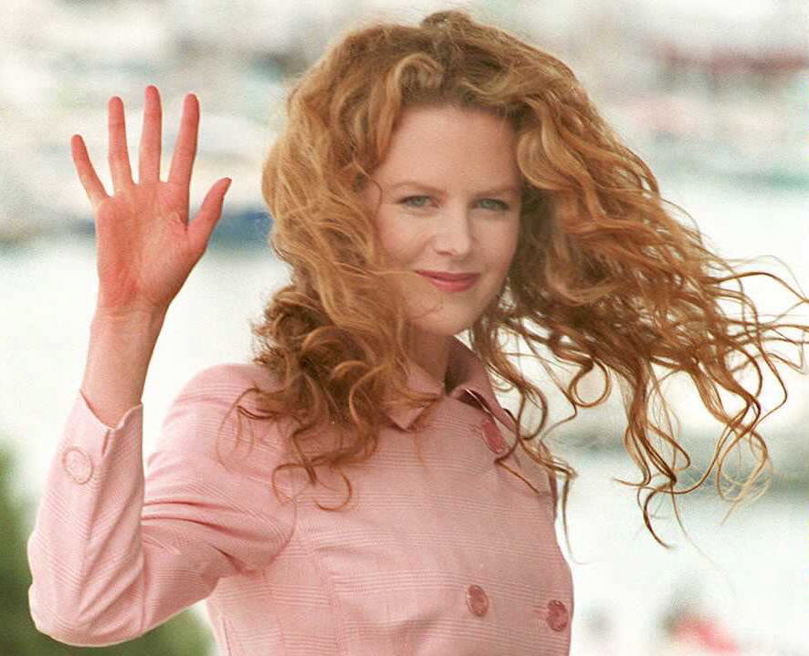 Nicole Kidman waves to photographers at the Cannes Film Festival during the press presentation of "To Die For." Her voluminous red curls cascade freely, complementing a soft pink coat and a bright, poised expression.