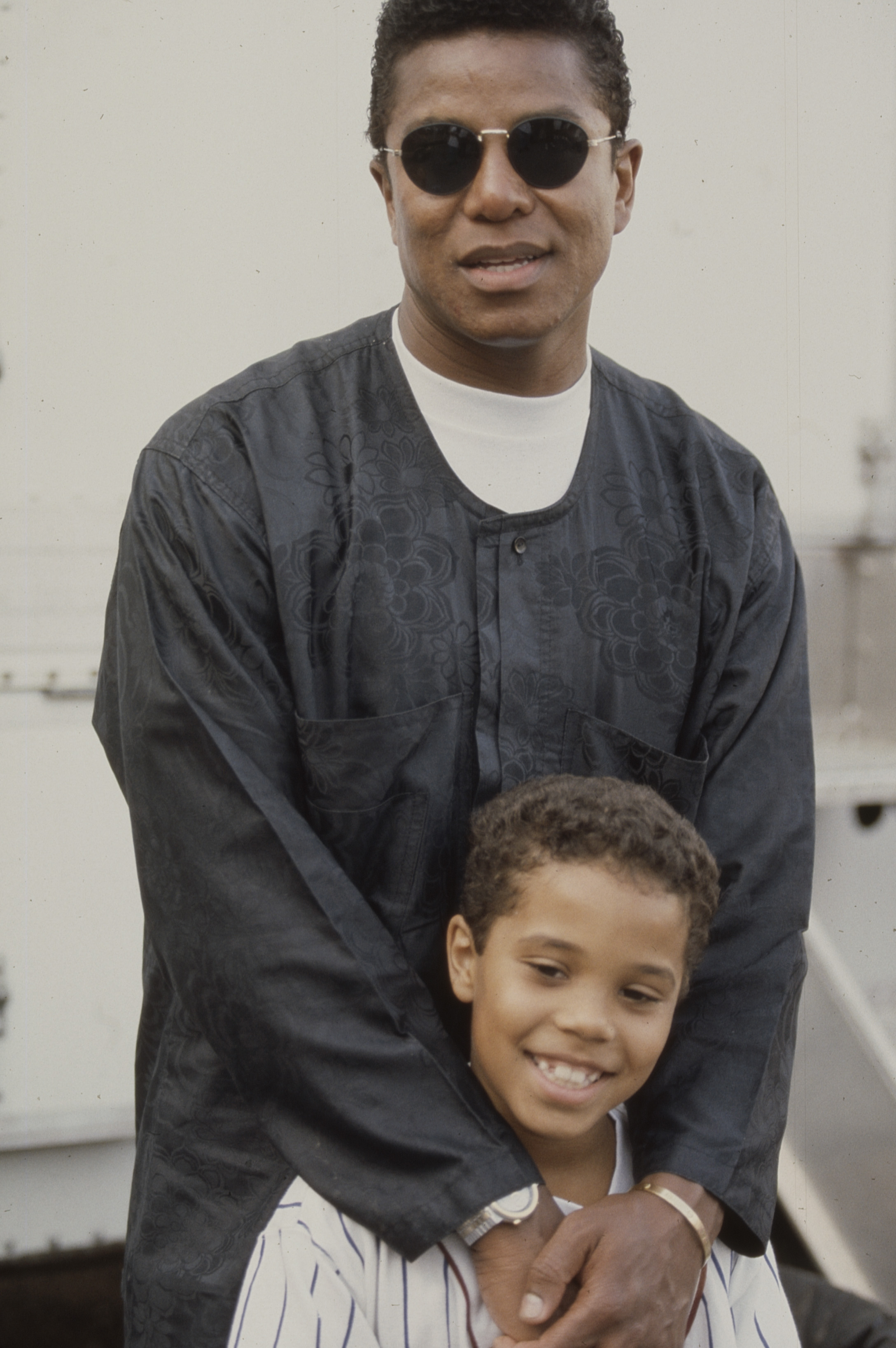Jermaine Jackson stands behind a young Alex Burrall, wrapping an arm around him in a warm, behind-the-scenes moment from The Jacksons: An American Dream. Dressed in a relaxed, patterned top and sunglasses, he blends star presence with a protective, almost fatherly ease.