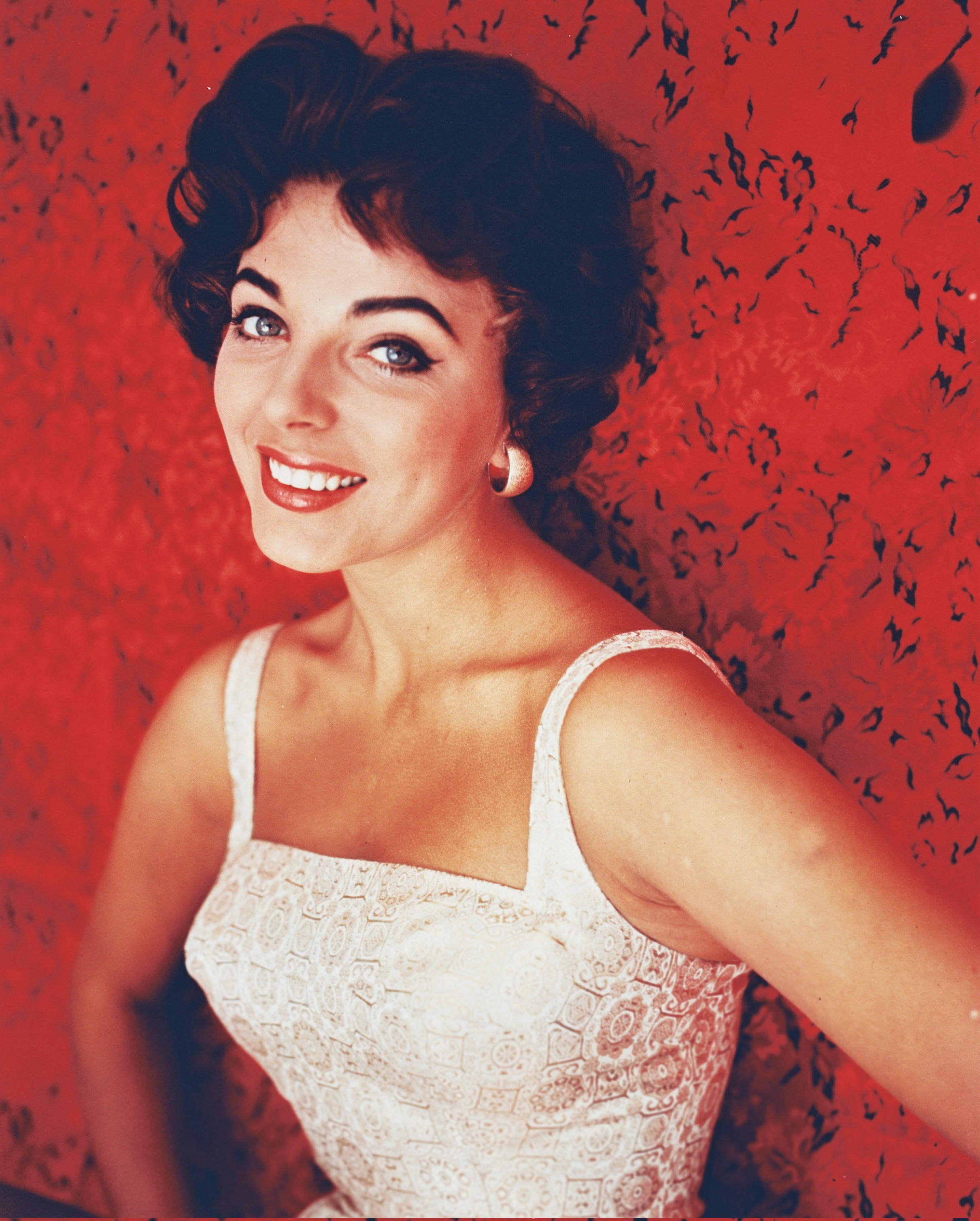 Joan Collins smiles brightly in a studio portrait set against a vivid red backdrop. She wears a sleeveless white brocade top with a square neckline, her hair styled in soft dark curls and her makeup featuring winged eyeliner and a classic red lip.