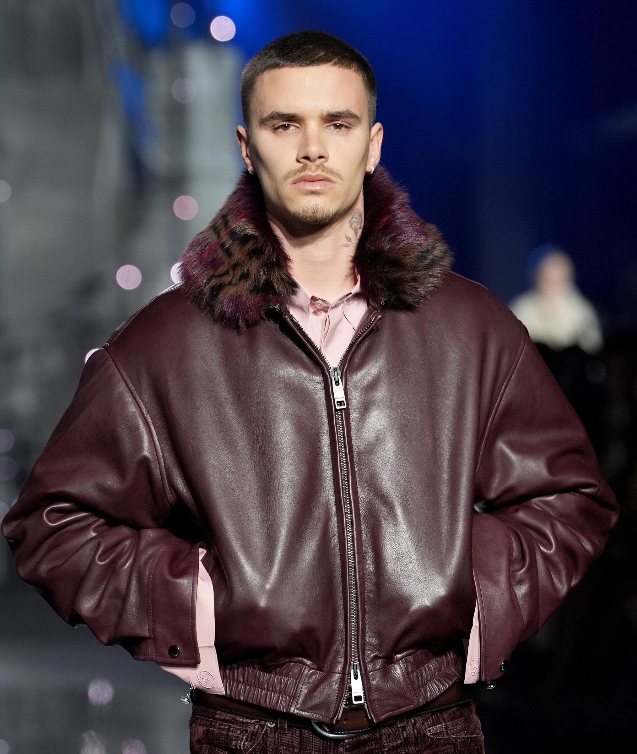 Romeo Beckham on the Burberry catwalk at London Fashion Week, February 2026 — buzz cut, a neck tattoo, and a burgundy leather bomber jacket with a leopard-print fur collar, his jawline a direct echo of his father's.