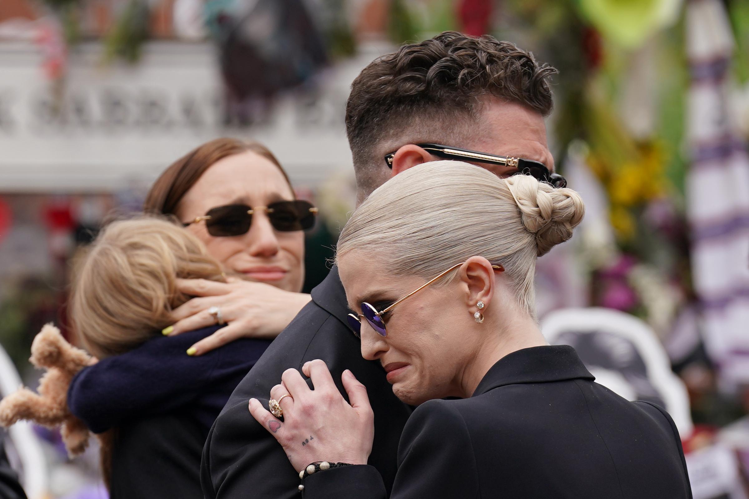 Jack and Kelly Osbourne share a tight embrace as they stand before the handwritten notes and floral tributes left at the Black Sabbath Bench and Bridge on Broad Street in Birmingham on July 30, 2025. With mourners gathered nearby, the siblings appear visibly overcome as they quietly reflect on the loss of their father, Ozzy Osbourne, whose body was returned to his home city.