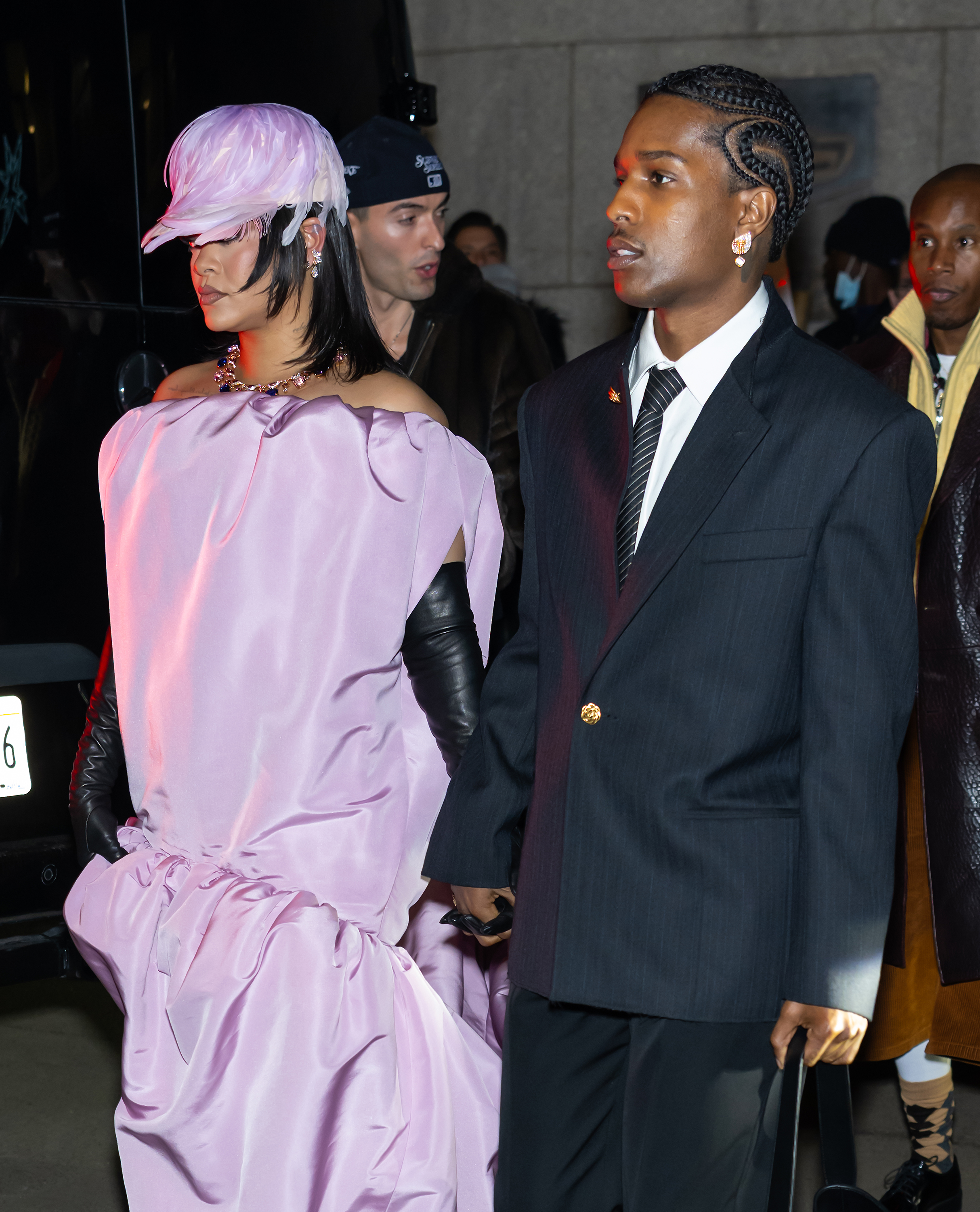Captured mid-stride, Rihanna and A$AP Rocky walk hand in hand through a crowd. She clutches her gown while he leads with focus, both looking composed and stylish in motion.