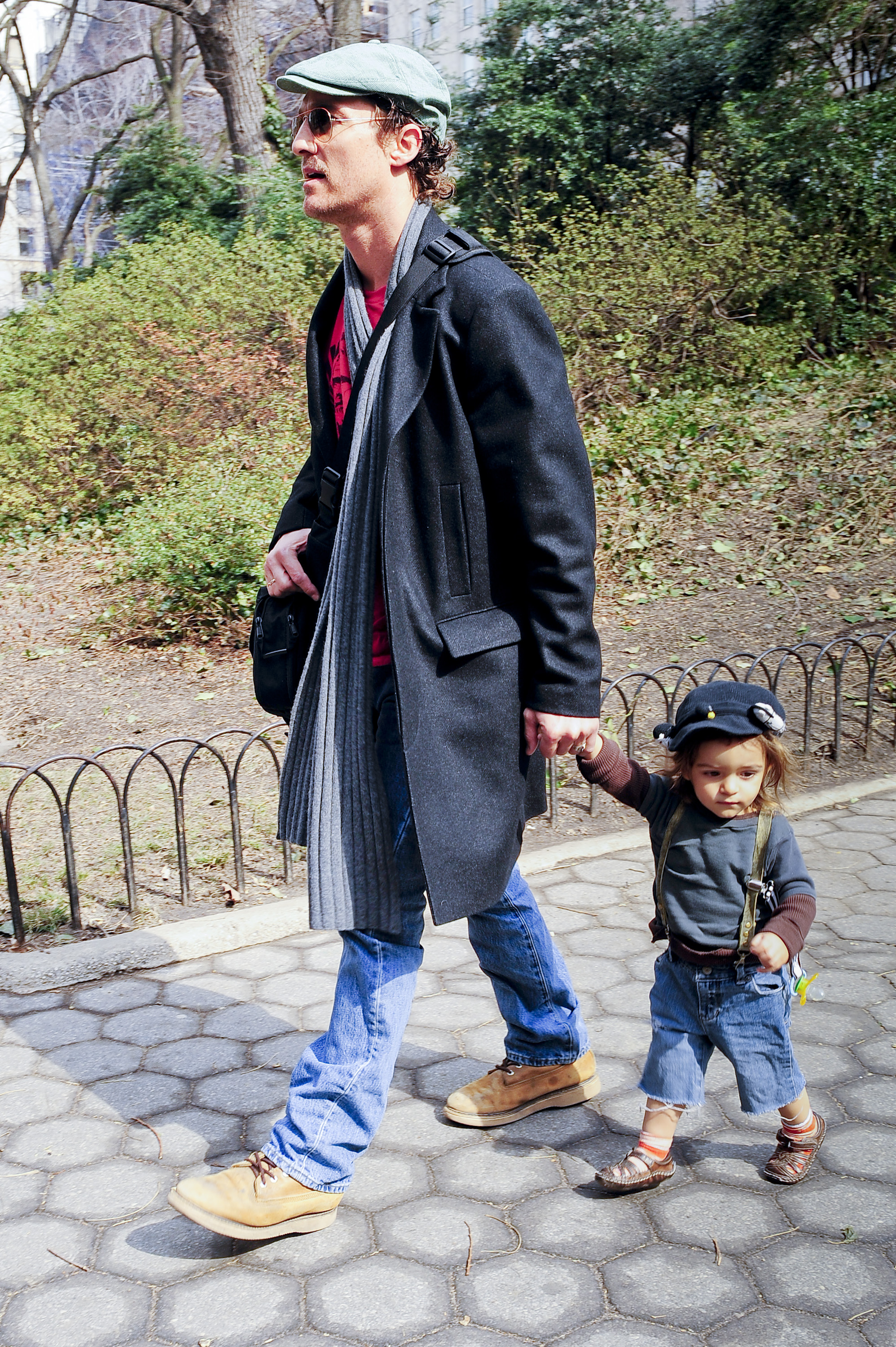 Levi McConaughey walks while holding Matthew McConaughey’s hand. He wears a small cap, long-sleeve top, shorts, and suspenders. Matthew McConaughey is dressed in a long coat, scarf, jeans, and a cap.