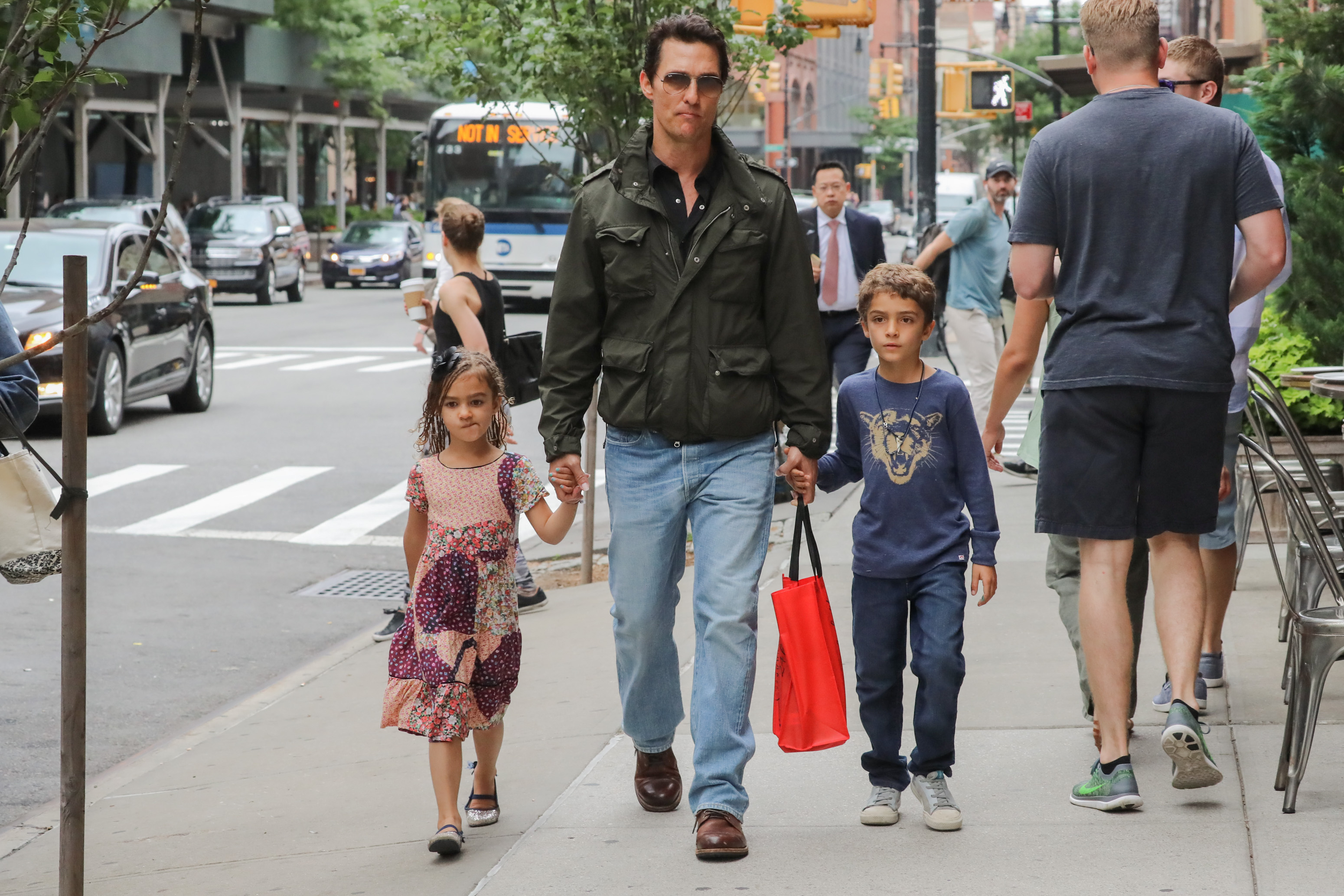 Vida McConaughey and Levi McConaughey walk on either side of Matthew McConaughey, each holding his hands. Vida wears a patterned dress while Levi wears a long-sleeve top and pants. They walk together along a city sidewalk with Matthew in a jacket and jeans.