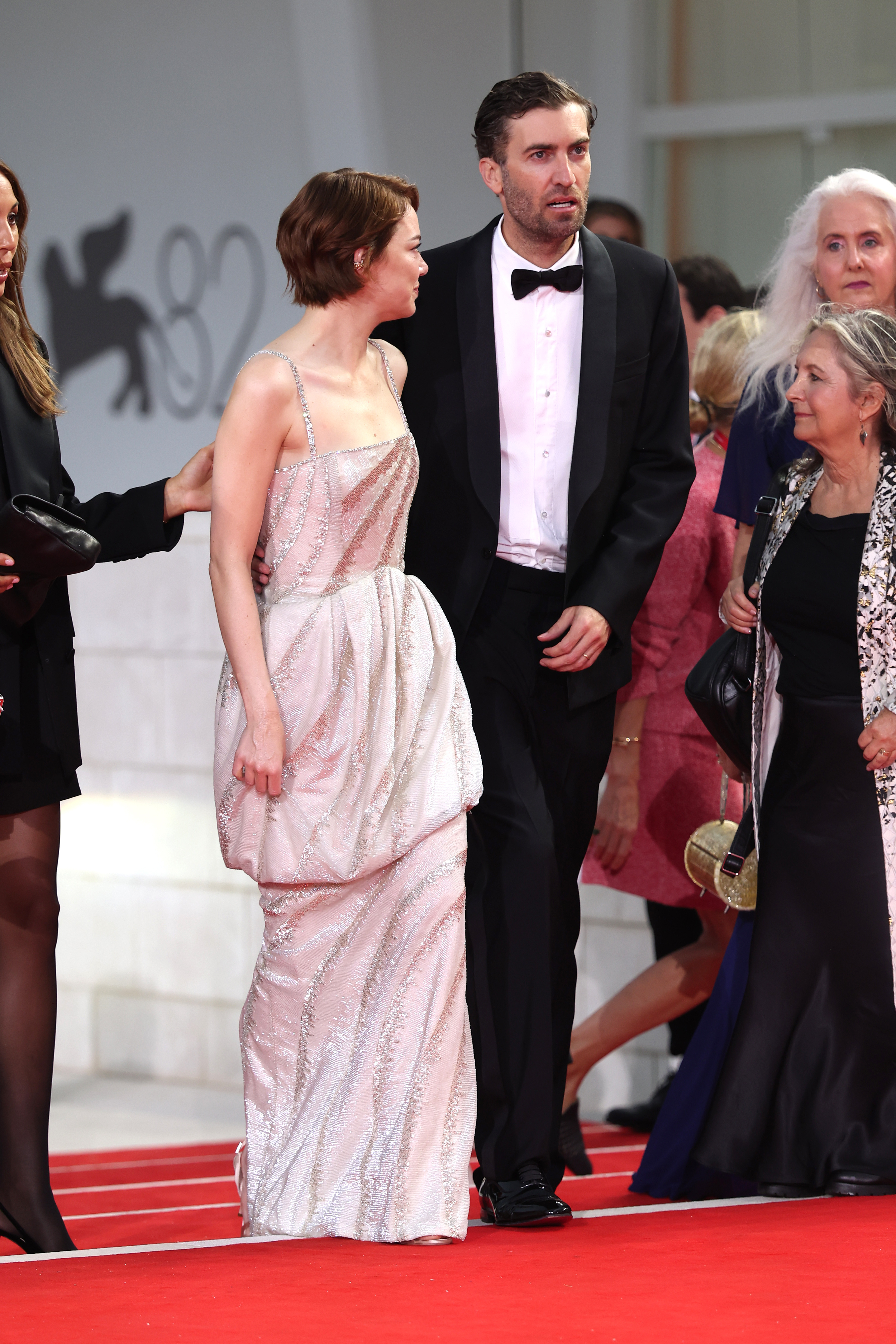 Emma Stone stood arm-in-arm with her husband, Dave McCary, wearing a shimmering silver gown with a sculptural peplum silhouette and delicate beaded straps. Her cropped hairstyle, still in early regrowth, offered a bold contrast to the romantic design.