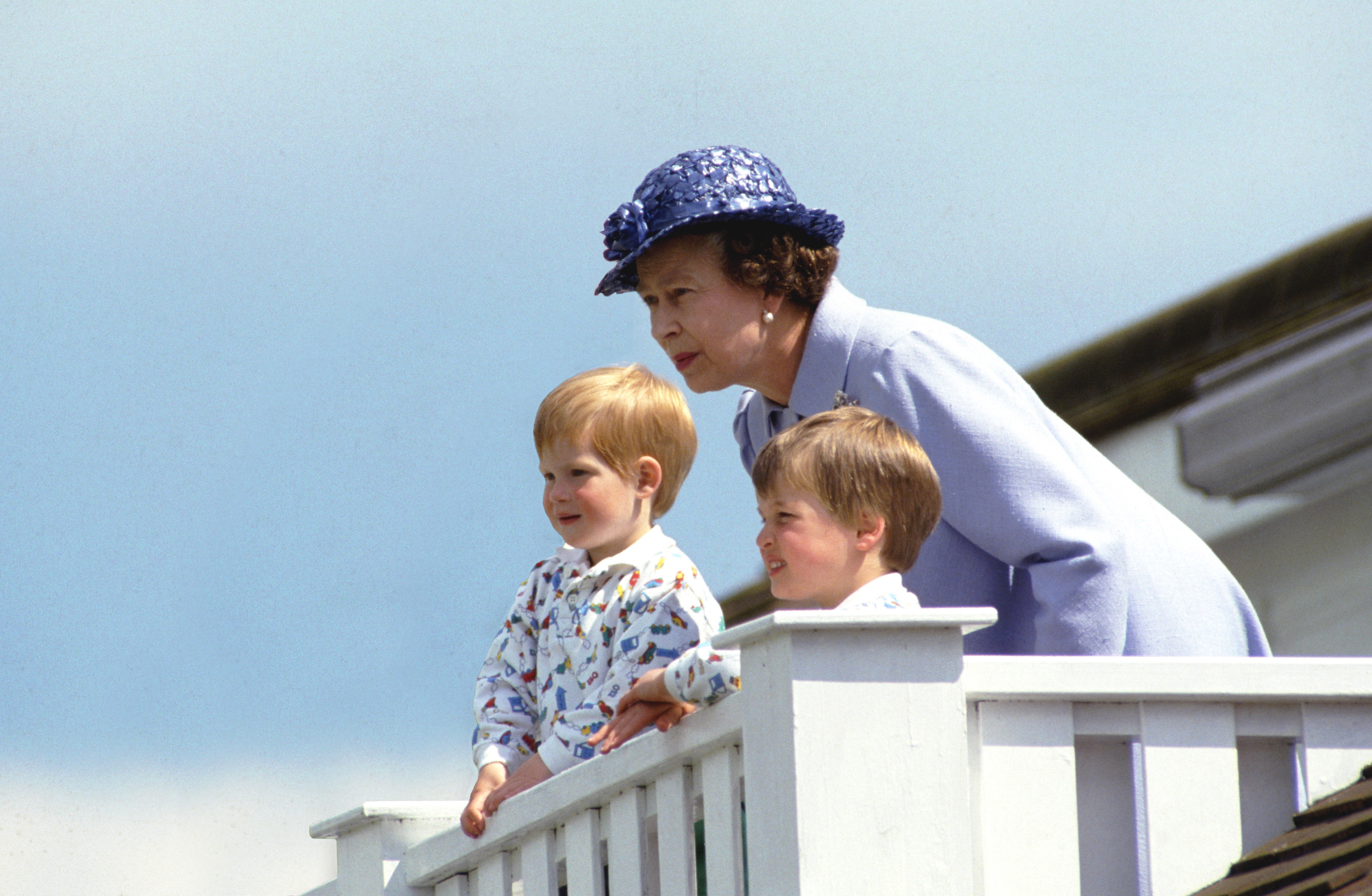 The late Queen Elizabeth II was pictured leaning in attentively beside a young Prince William and Prince Harry as they looked out from the Royal Box at Guards Polo Club in Windsor on 14 June 1987. Dressed in a soft blue ensemble and matching hat, the monarch appeared engaged and protective, while the two young princes — one fair-haired and the other slightly older — stood side by side.