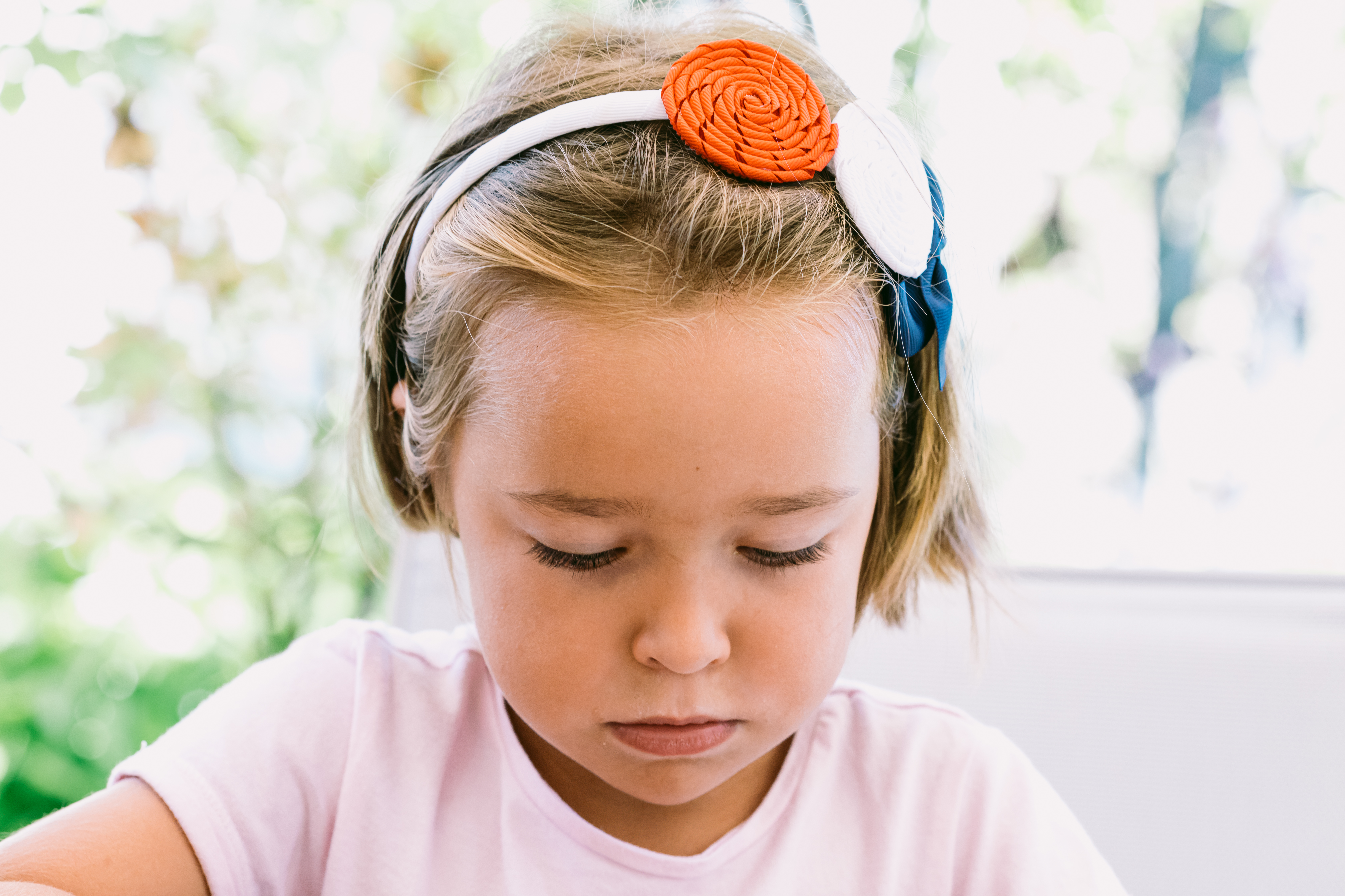 Portrait of a serious blonde-haired little girl, wearing a white headband with blue, red and white decorations, with a garden in the background | Source: Getty Images