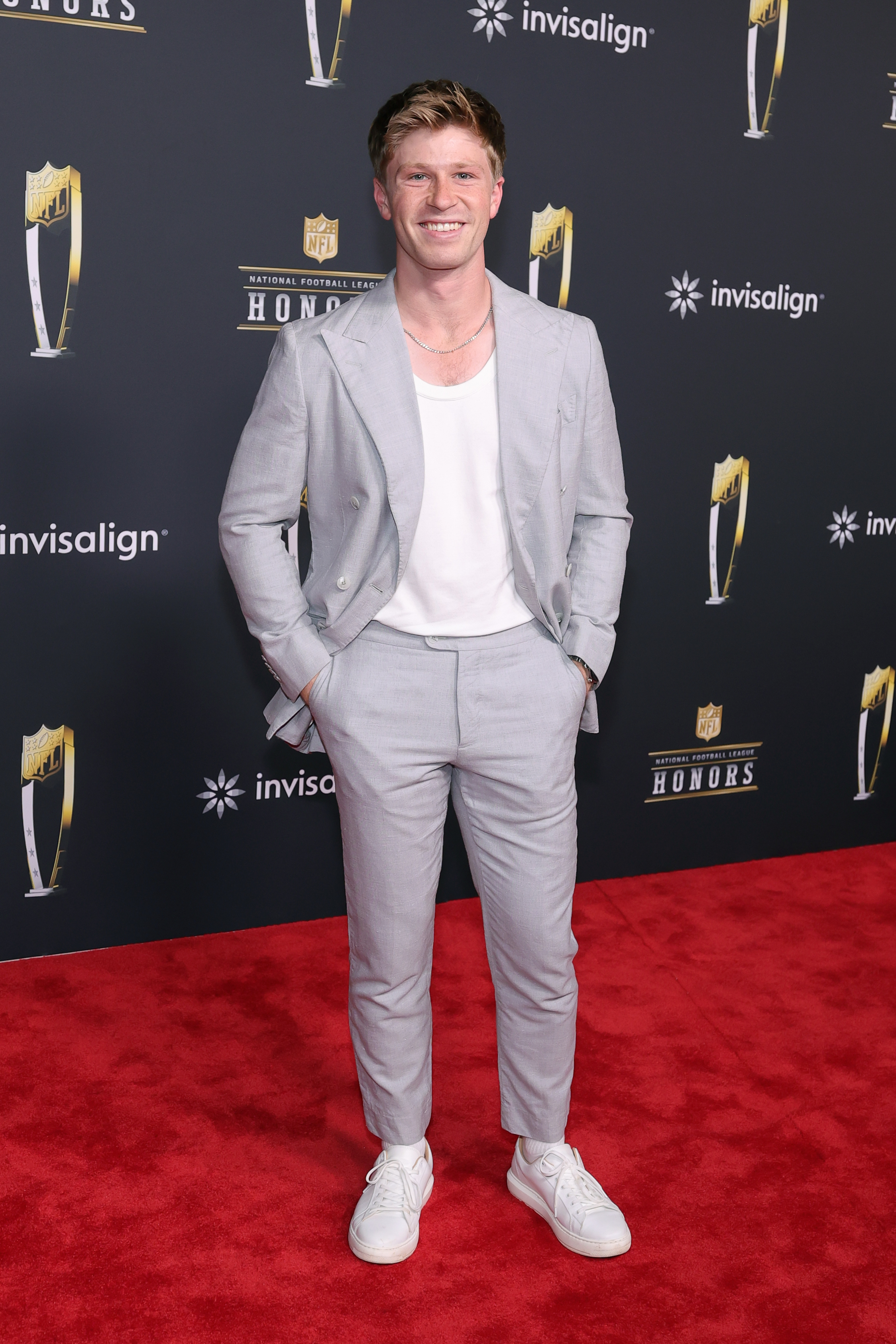Robert Irwin smiles brightly in a light gray suit paired with a white T-shirt and sneakers, blending youthful charm with modern red-carpet style.