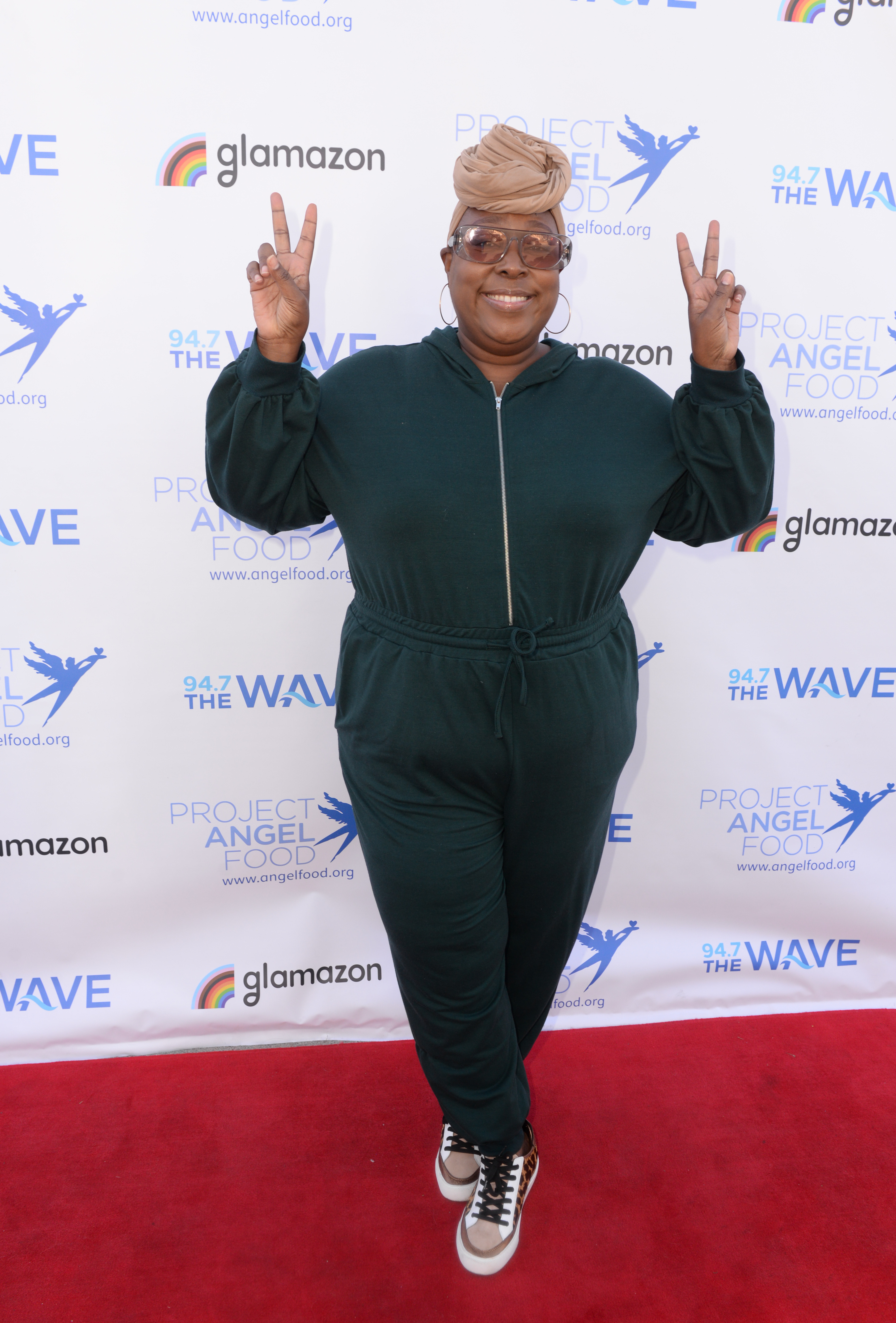 Loni Love joins Project Angel Food to deliver 2,000 Thanksgiving meals on November 25, dressed casually in a dark jumpsuit, sneakers, glasses, and a head wrap while flashing peace signs.