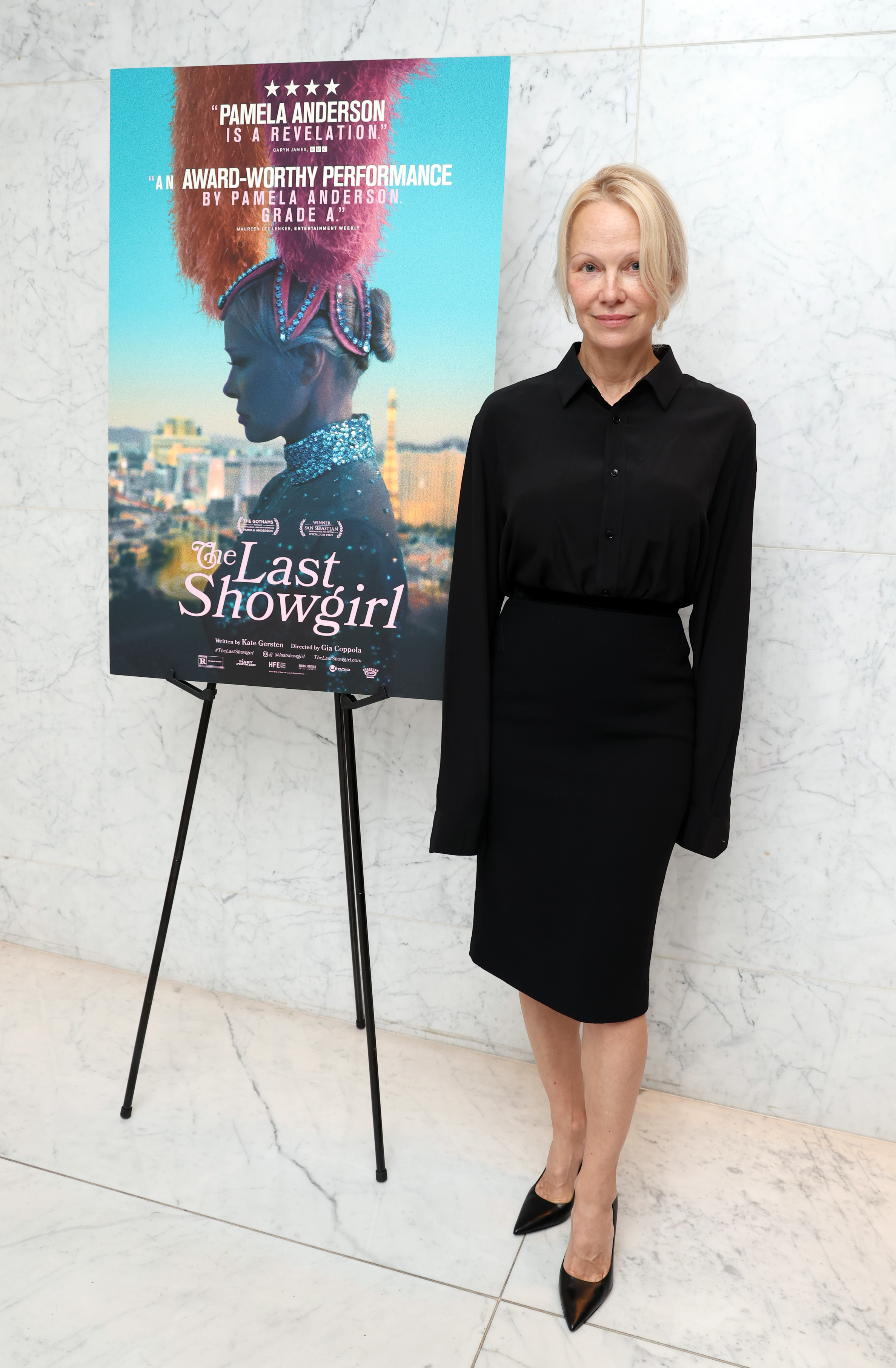 Pamela Anderson poses beside a promotional poster for The Last Showgirl during a special screening hosted by Roadside Attractions at CAA on December 7, 2024, in Los Angeles, California. She wears an all-black ensemble featuring a button-down blouse and knee-length skirt.