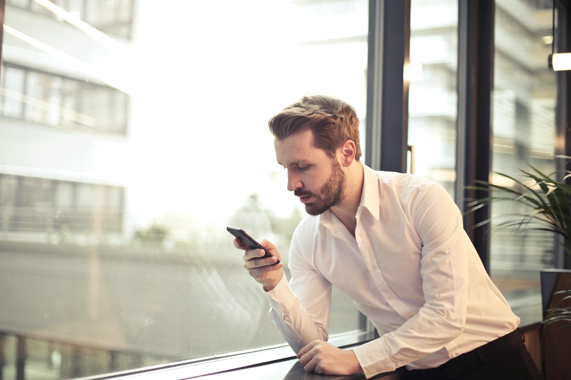 Hombre de pie cerca de una ventana mirando su teléfono | Fuente: Pexels