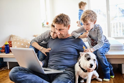 Father using laptop at home with children watching.| Photo: Getty Images.