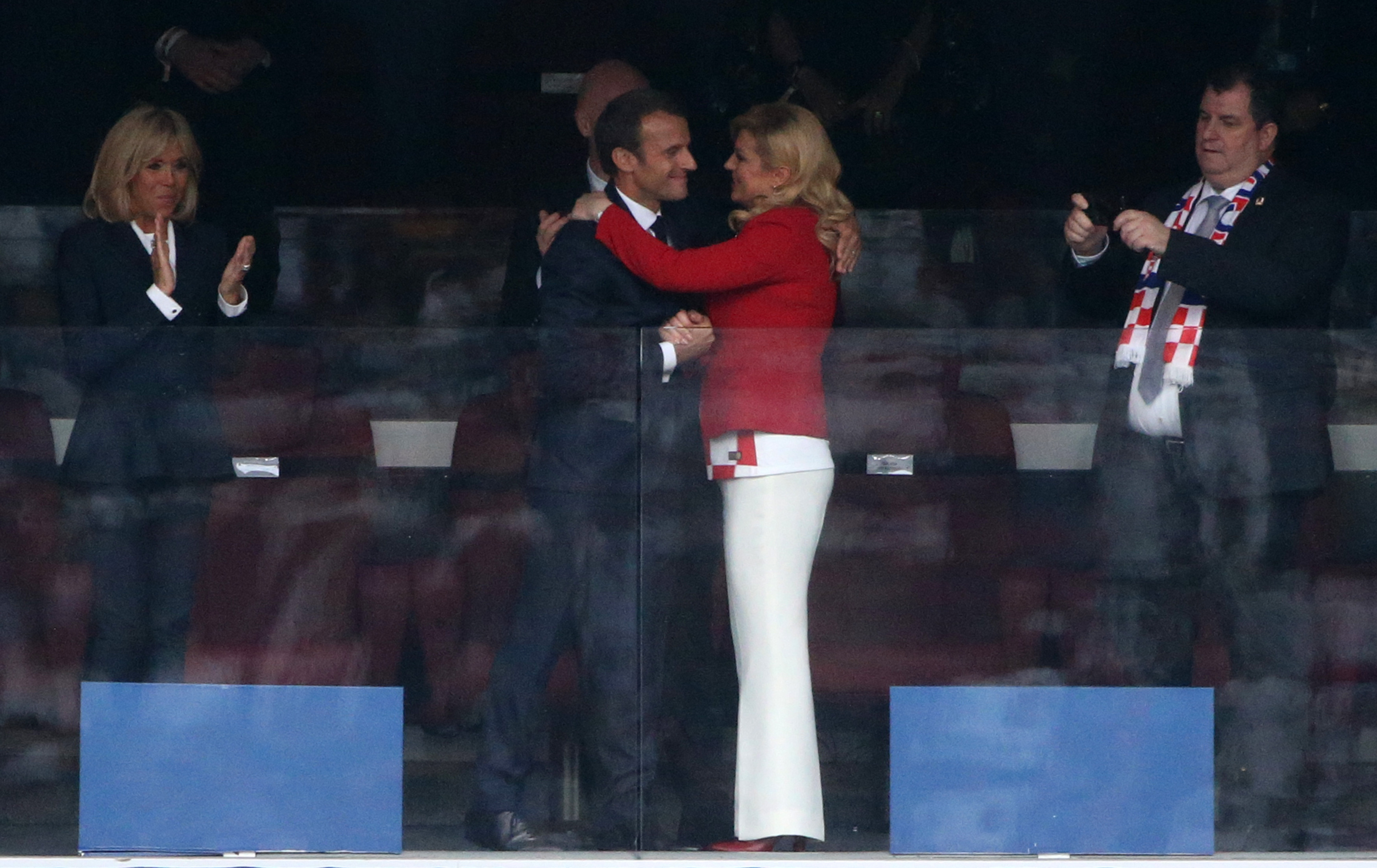 In another moment, Emmanuel Macron wraps both arms around Kolinda Grabar-Kitarović in an embrace, while Brigitte Macron stands nearby clapping, observing the scene.