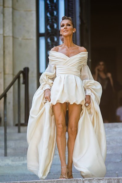  Celine Dion wears earrings, rings, a lustrous ivory-color off-shoulder drape mini dress with long flowing puff sleeves, anklets, shiny golden strappy heeled sandals, outside Alexandre Vauthier, during Paris Fashion Week -Haute Couture Fall/Winter 2019/2020, on July 02, 2019 in Paris, France | Photo: Getty Images