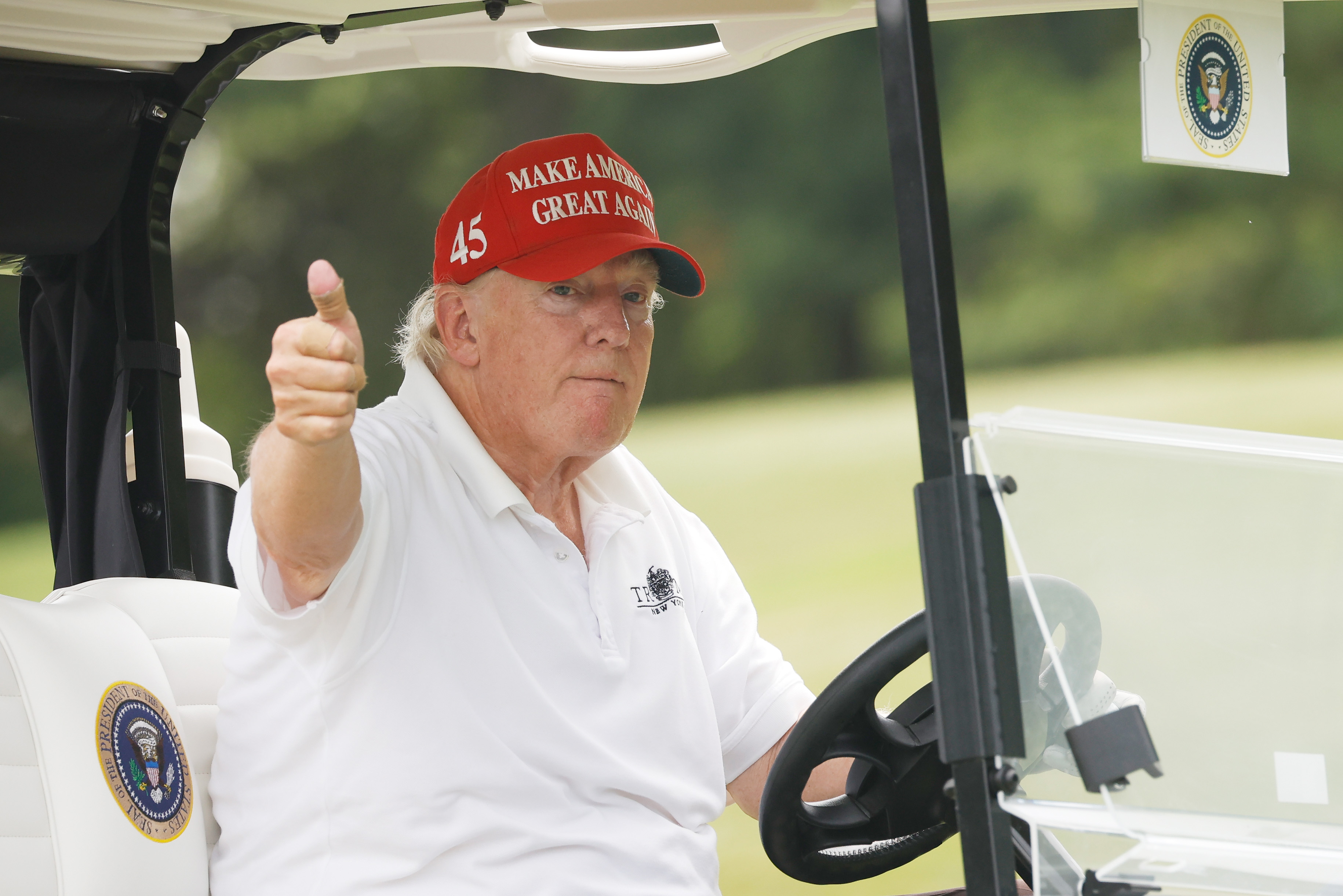 Seated in a golf cart, Donald Trump gives a thumbs-up with a serious look on his face. The color difference between his face and neck is visible under the daylight.