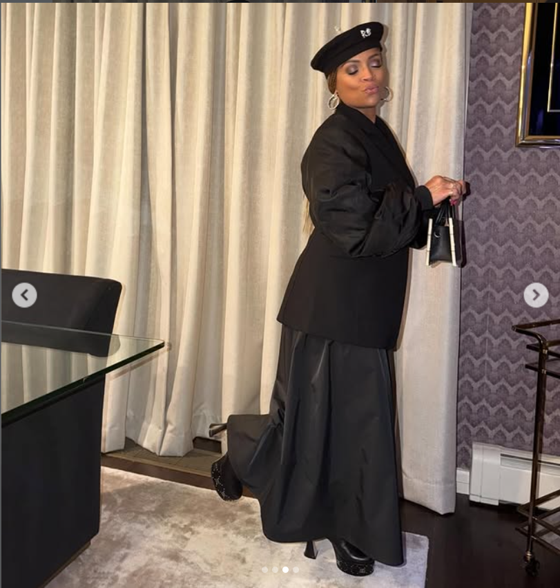 Simone Smith poses playfully indoors in a chic all-black ensemble, wearing a structured blazer, flowing skirt, and a beret, as she holds a small handbag and lifts one foot slightly. | Source: Instagram/sislovespurple