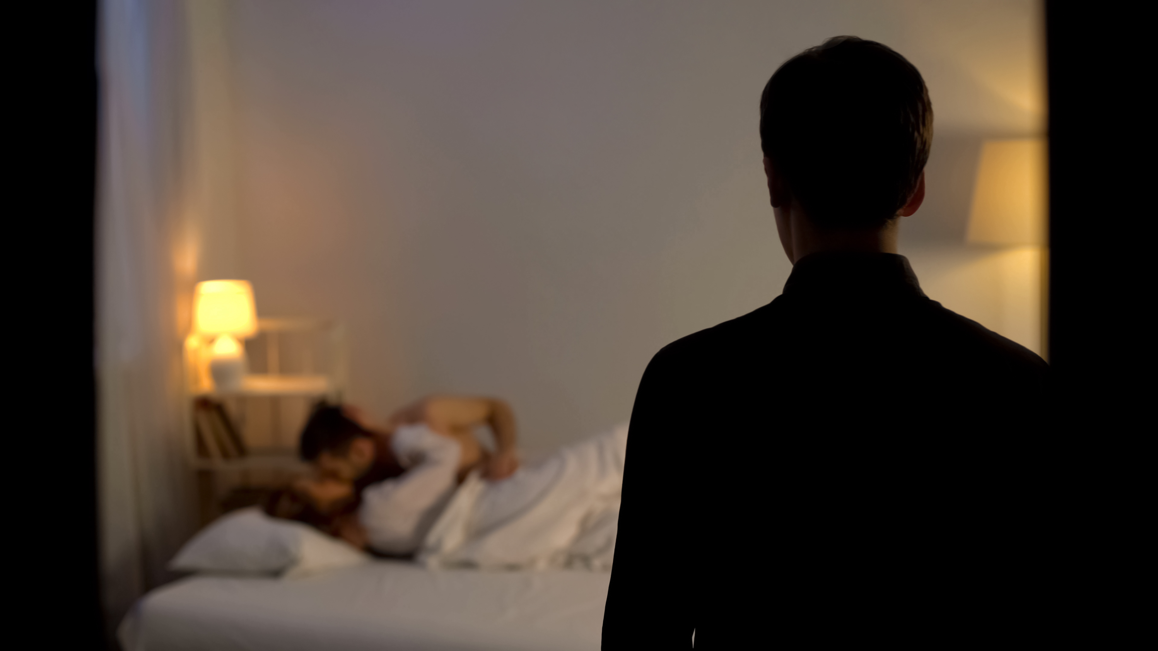 A husband stands in shock after catching his wife cheating with a lover in bed | Source: Shutterstock