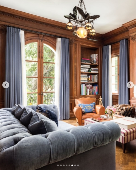 Floor-to-ceiling walnut panelling lines the walls of this study, where arched French doors draw in the garden light and a tufted slate-blue Chesterfield sofa dominates the foreground. A gothic-style bat chandelier fitted with amber bulbs hangs from the carved cornice — a nod to the Osbourne sensibility hiding quietly inside an otherwise classically furnished room. | Source: Instagram/top.music.songs