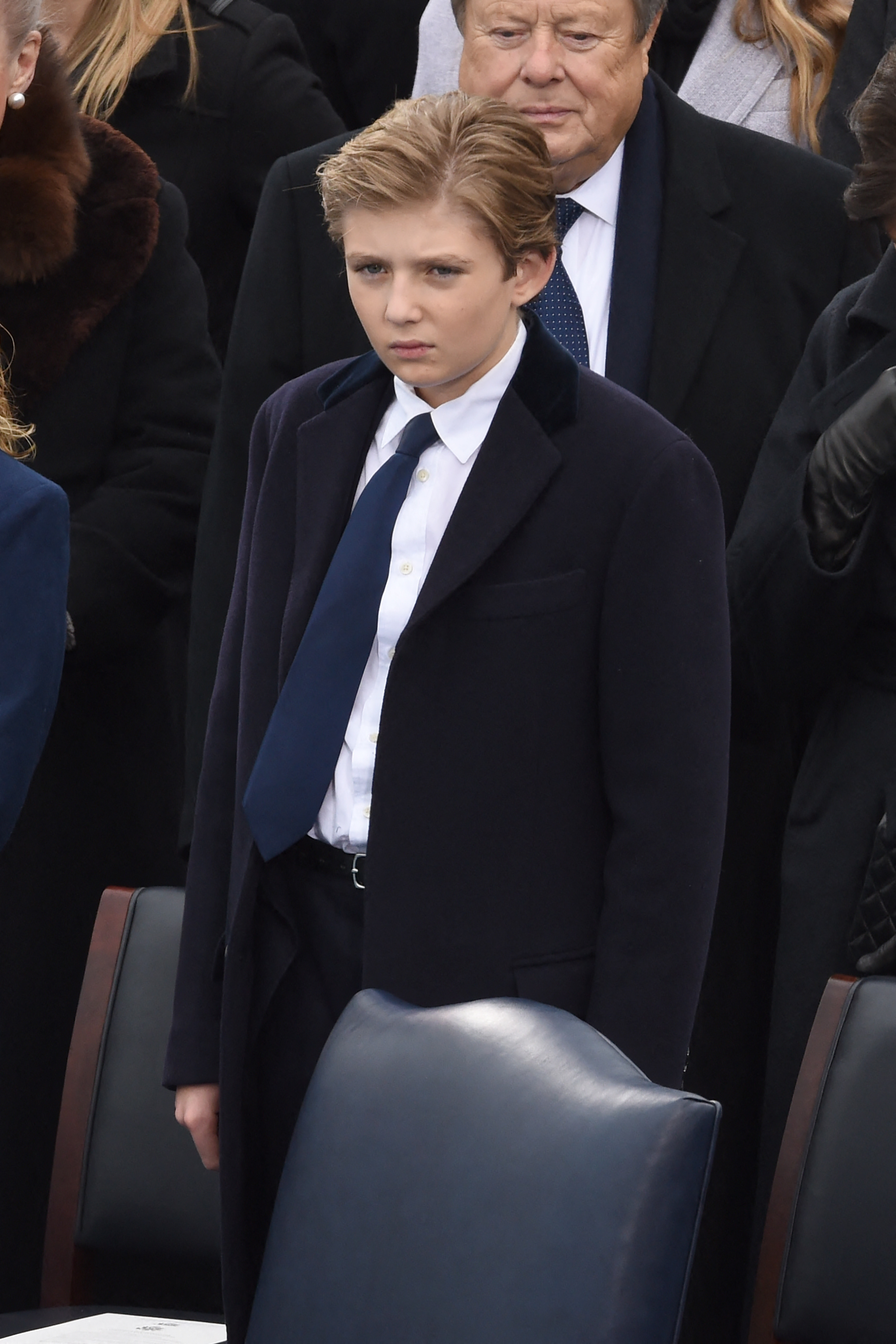 Barron Trump stands solemnly in a navy overcoat during the swearing-in ceremony at the U.S. Capitol. Dressed in a white shirt and blue tie, he appears focused amid the gathered crowd. The moment unfolds as Donald Trump is inaugurated as the 45th President of the United States.