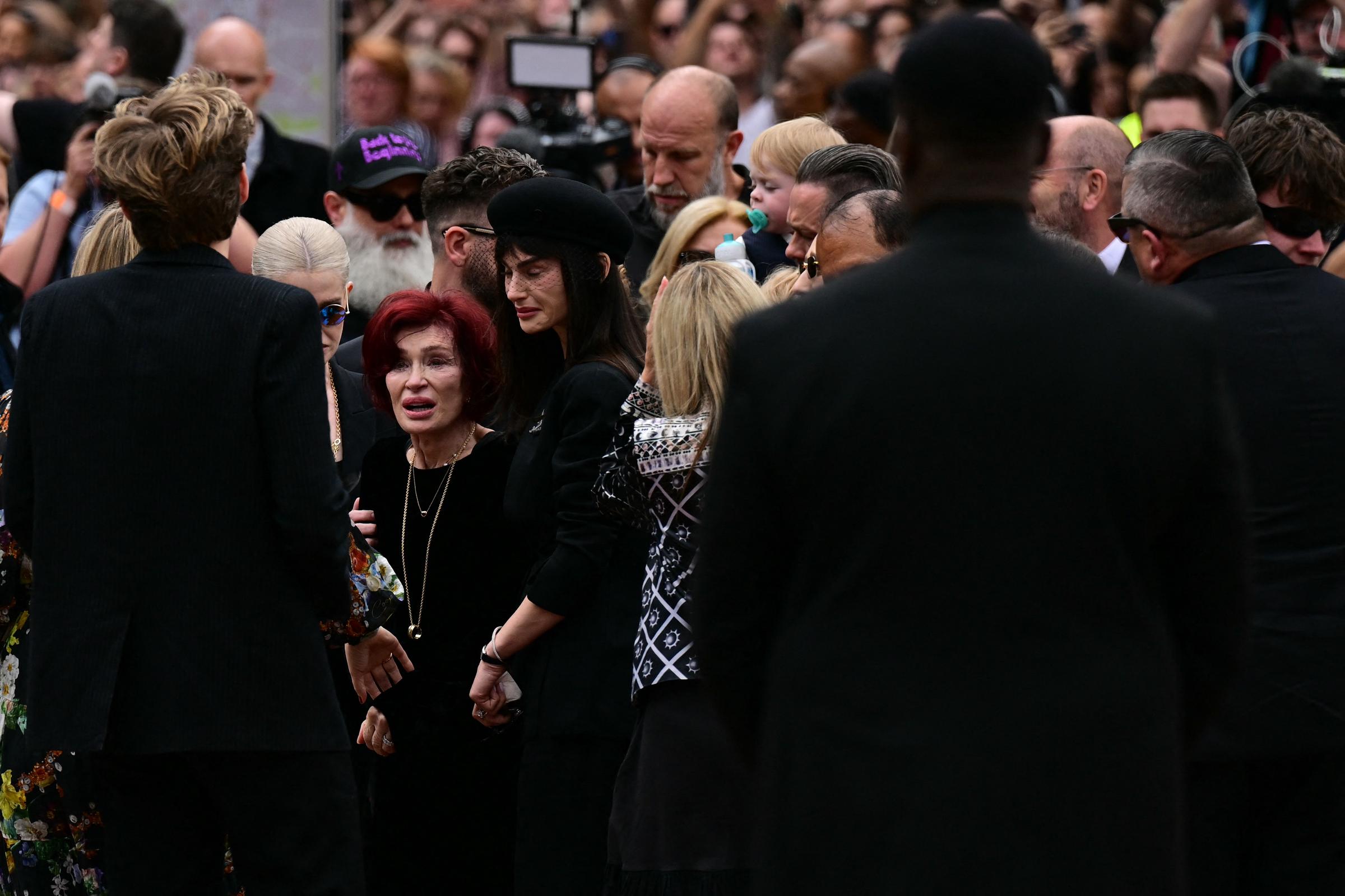 Sharon Osbourne stands visibly shaken, her face etched with grief as she's surrounded by mourners during Ozzy's funeral. The overwhelming emotion in the crowd mirrors the deep loss felt by his family and fans alike.