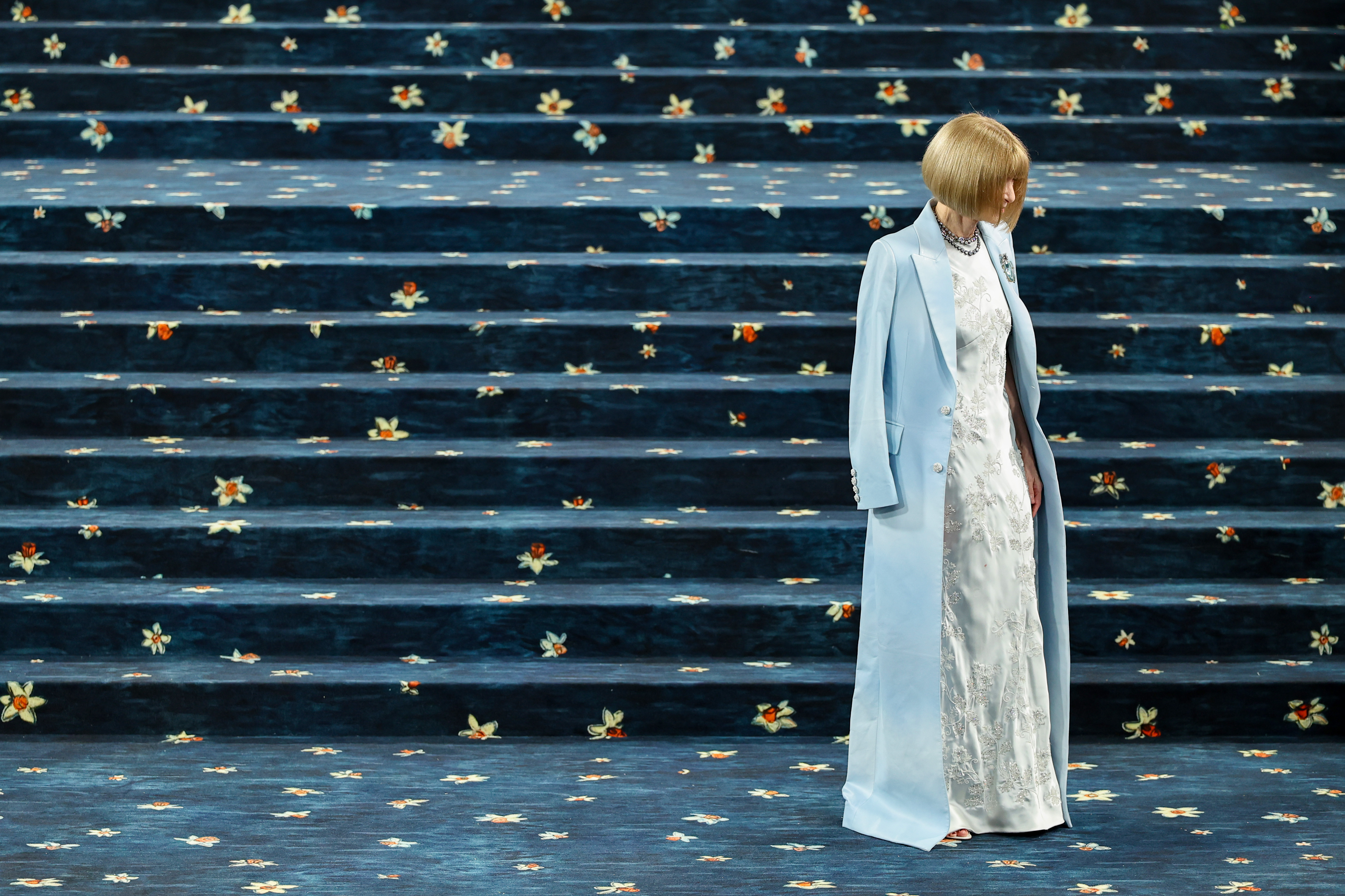 Anna Wintour pauses by the Met Gala staircase, dressed in an icy blue gown and baby blue blazer. The event celebrated "Superfine: Tailoring Black Style," spotlighting Black designers and cultural storytelling through fashion.