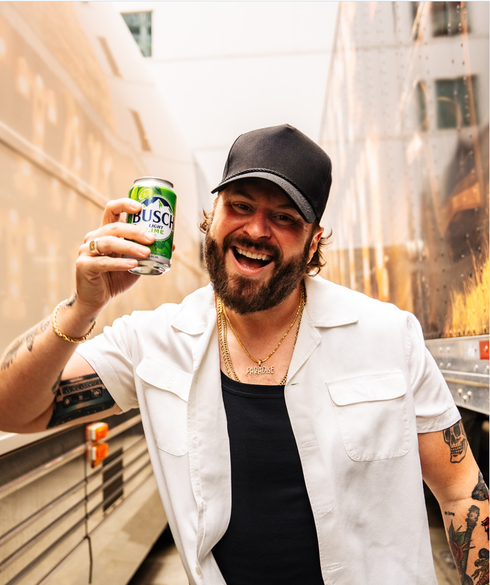 Nate Smith flashes a wide smile while raising a can of Busch Light Lime, posing backstage in a white short-sleeved shirt layered over a black top with his tattooed arms on display. | Source: Instagram/natesmith
