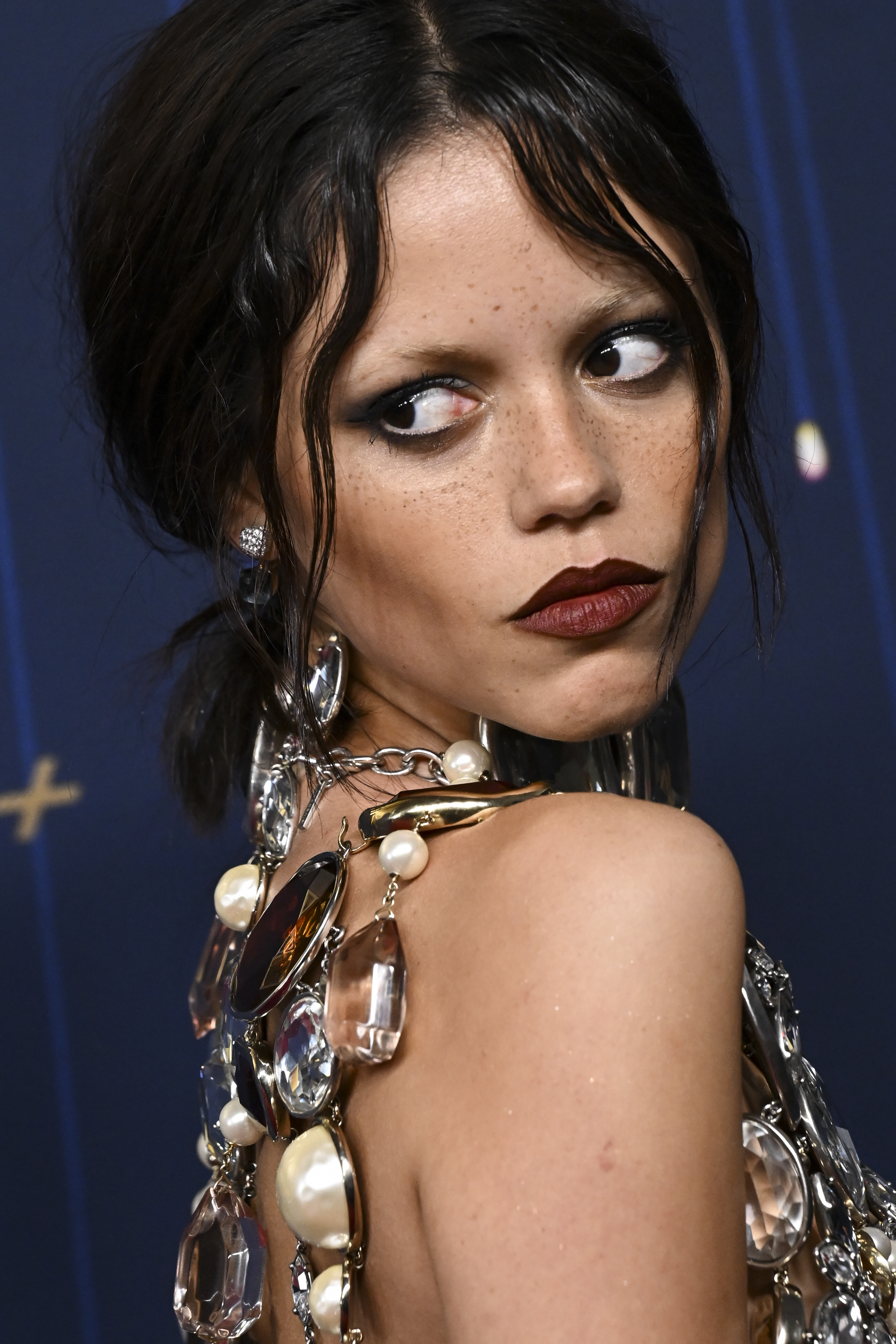 Jenna Ortega's facial appearance is captured at the 77th Primetime Emmy Awards in Los Angeles, California. She has on a dark maroon lipstick, and her black hair appears ruffled and parted in the middle.
