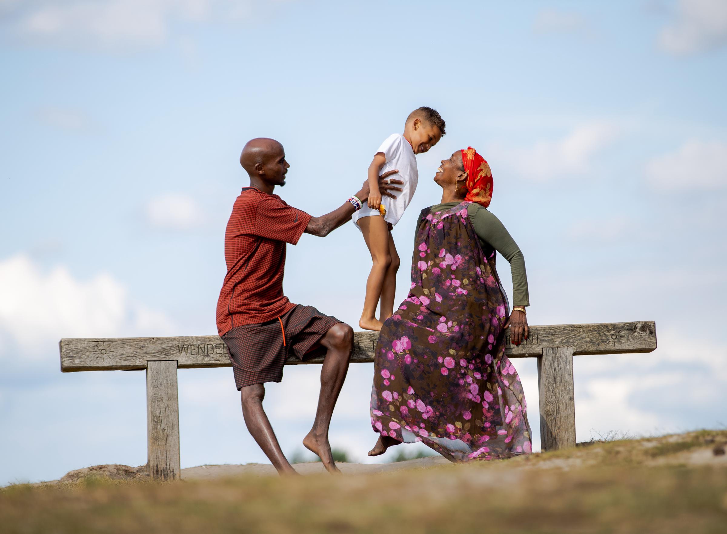 Sir Mo Farah sits on a wooden bench at Frensham Great Pond, Farnham, on 12 July 2019, holding his young son, Hussein, aloft between himself and his mother, Aisha, whose head is thrown back in laughter. It is a quietly extraordinary image — the little boy named Hussein, the same name Farah was born with and later had taken from him, now carried proudly by the next generation.