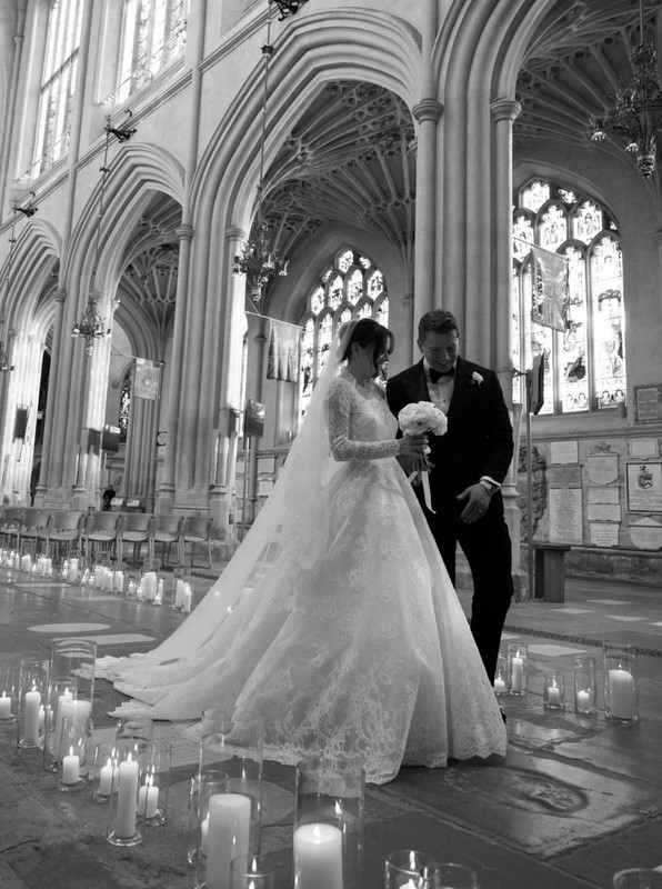Beneath the vaulted arches of Bath Abbey, Holly Ramsay and Adam Peaty shared a quiet moment surrounded by flickering candlelight. Holly's dramatic train swept across the stone aisle, while Adam stood close beside her in a classic tuxedo. The black-and-white frame captured the architectural beauty of the setting and the intimacy of the couple's exchange, just moments before or after their vows. | Source: Instagram/britishvogue