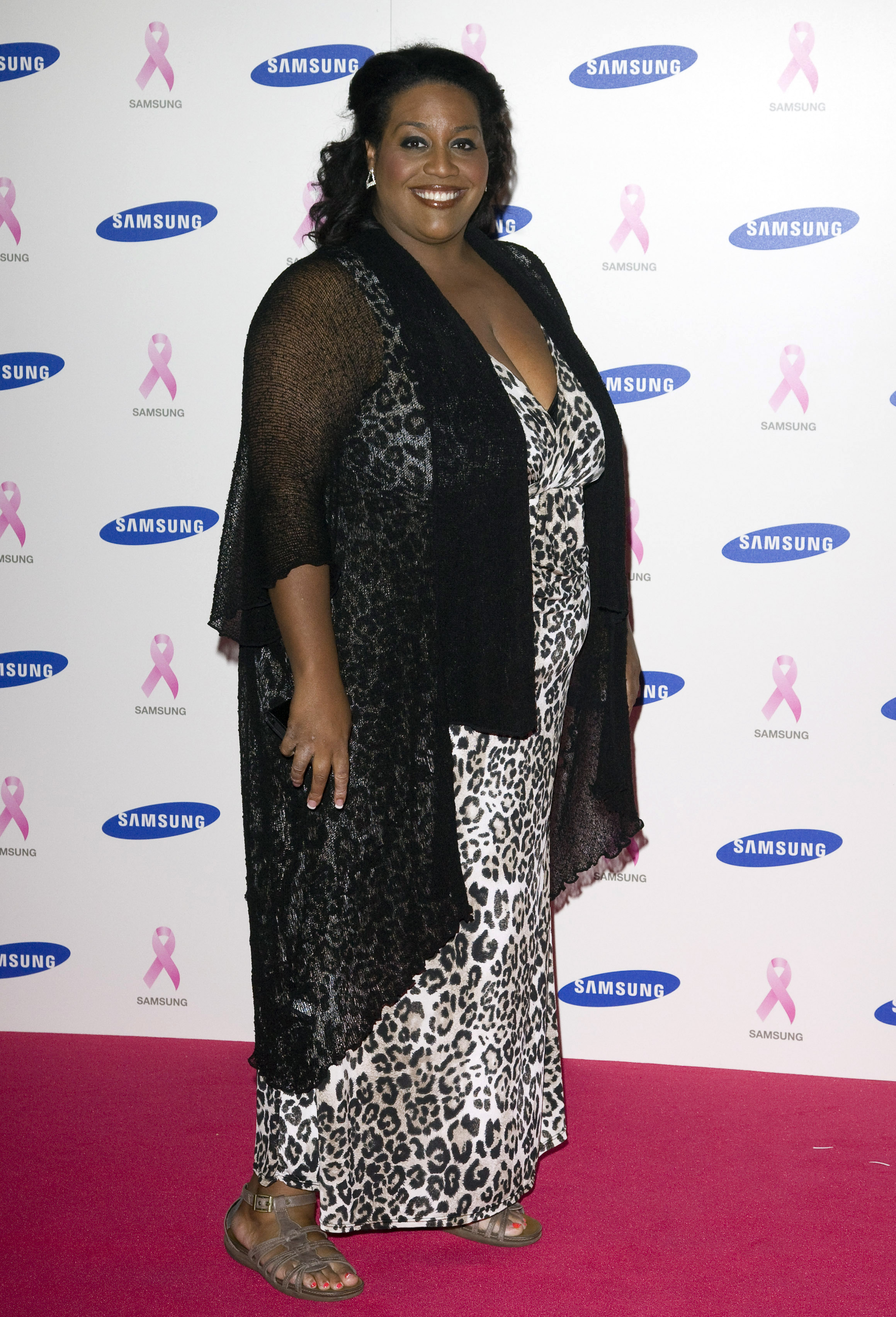 Striking a pose on the pink carpet, Hammond smiles for the cameras at the Samsung Pink Ribbon Celebration at the Royal Exchange, London, on 14 October 2010. She wears a leopard-print maxi dress and sheer black shawl, with sandals and delicate jewellery, surrounded by pink ribbon emblems and Samsung logos.